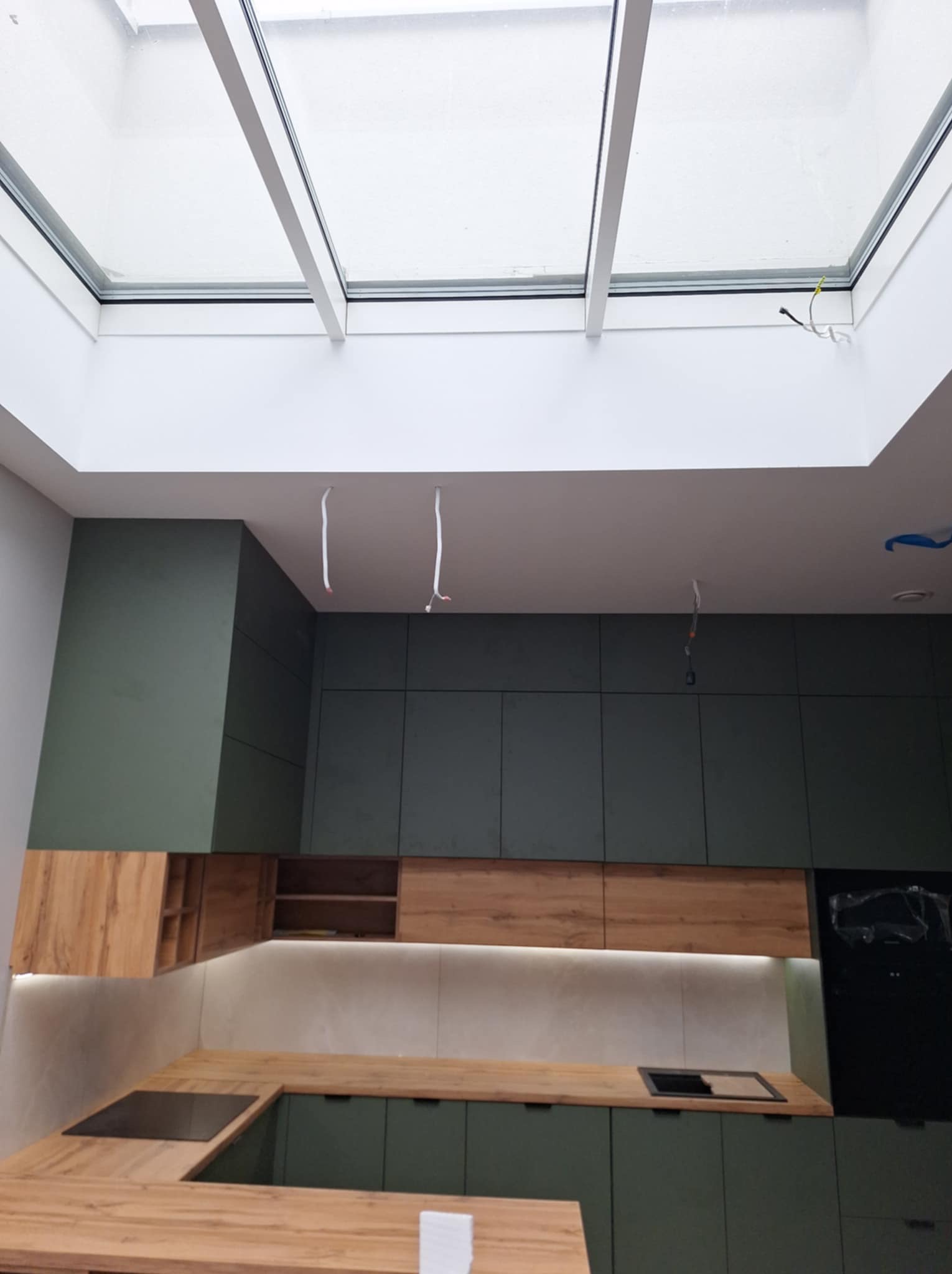 Kątowe ujęcie kuchni w trakcie montażu z zielonymi szafkami i drewnianymi blatami, widoczne niedokończone podłączenia elektryczne i okno dachowe.