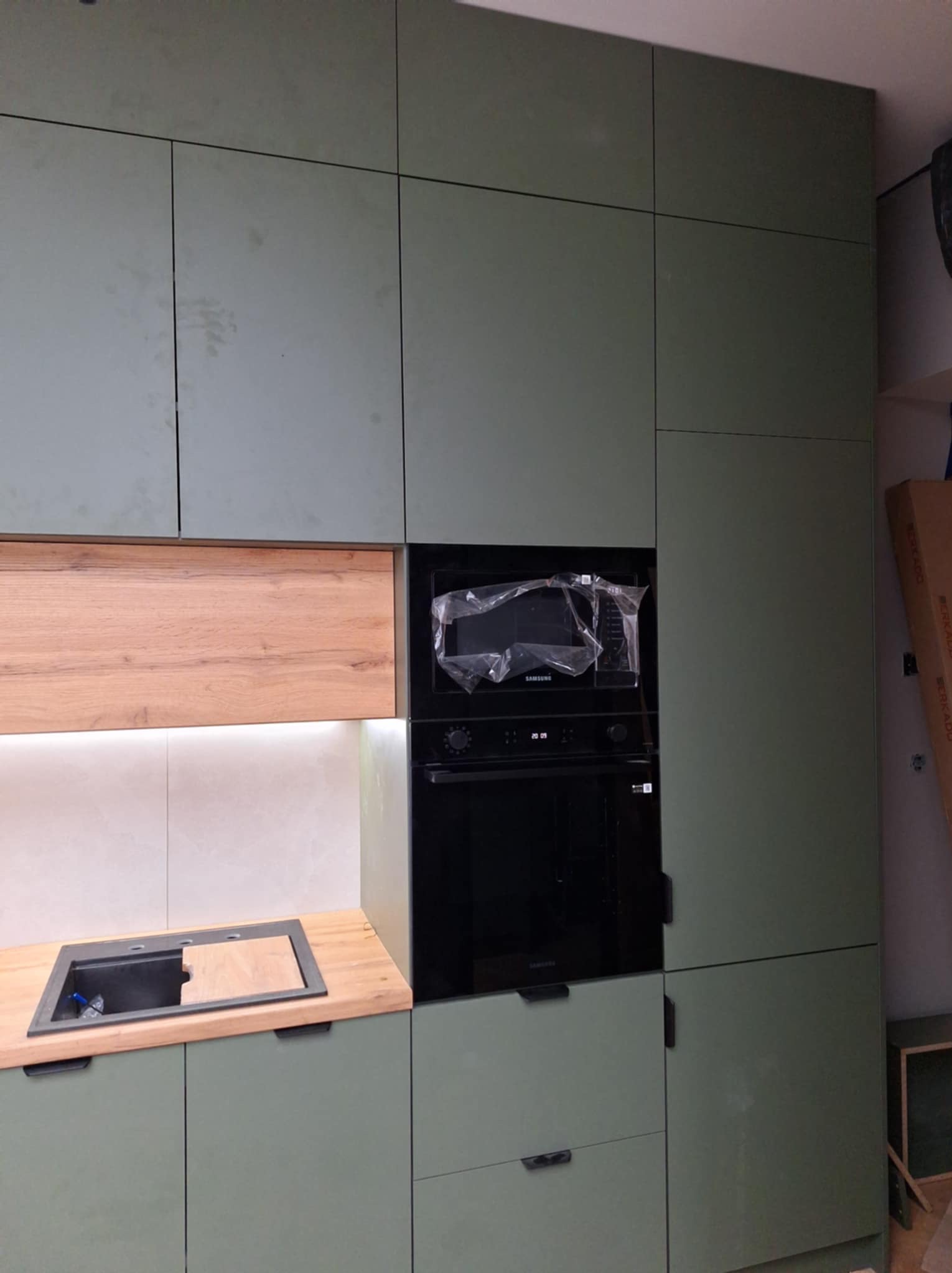 Fragment nowej kuchni z zielonymi frontami, drewnianym blatem i wbudowanym piekarnikiem z mikrofalówką, jeszcze w folii ochronnej.