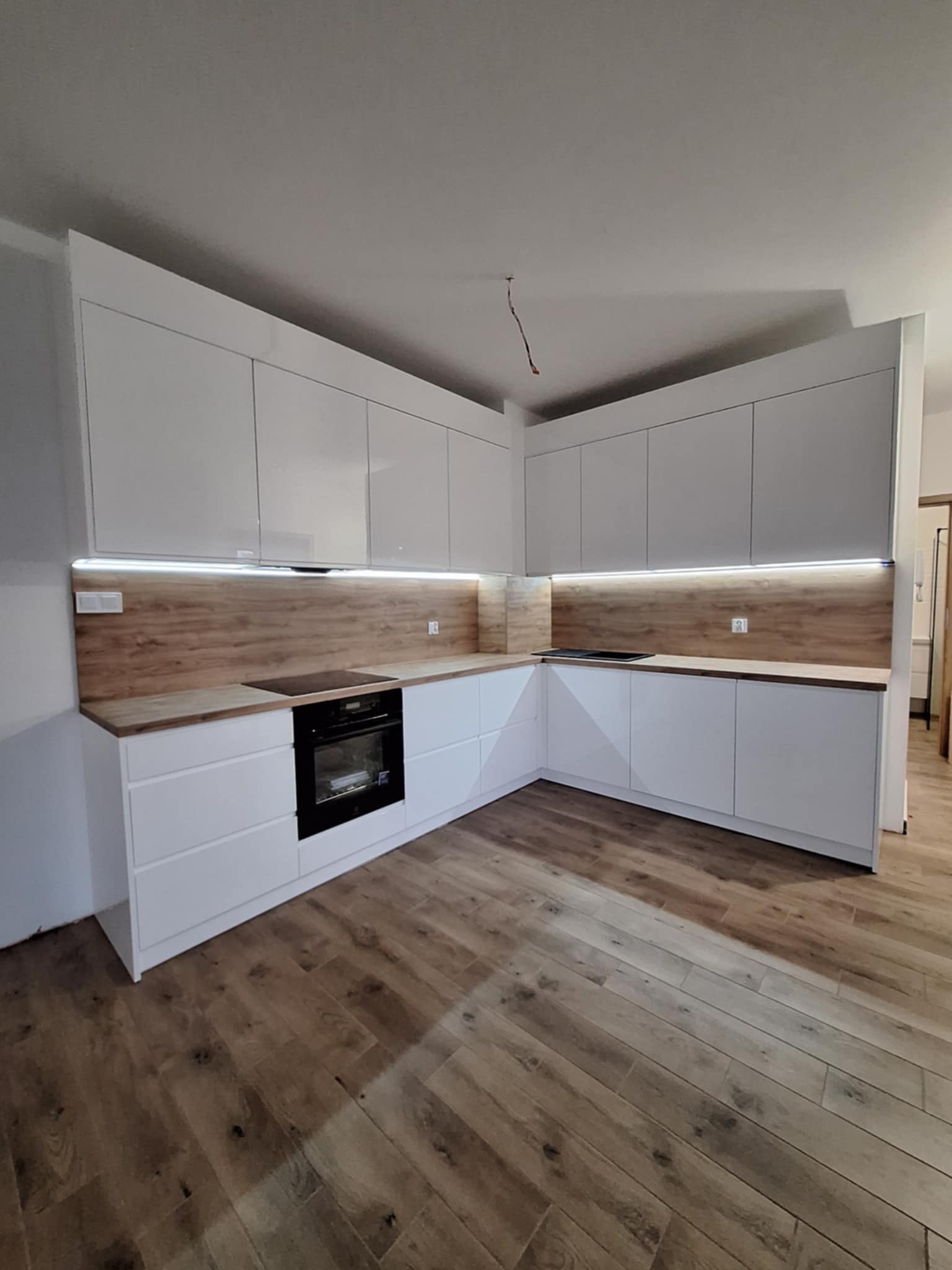 Nowoczesna, biała zabudowa kuchenna z drewnianym panelem nad blatem i oświetleniem LED, widok na narożnik kuchni z płytą grzewczą i piekarnikiem, drewniana podłoga.