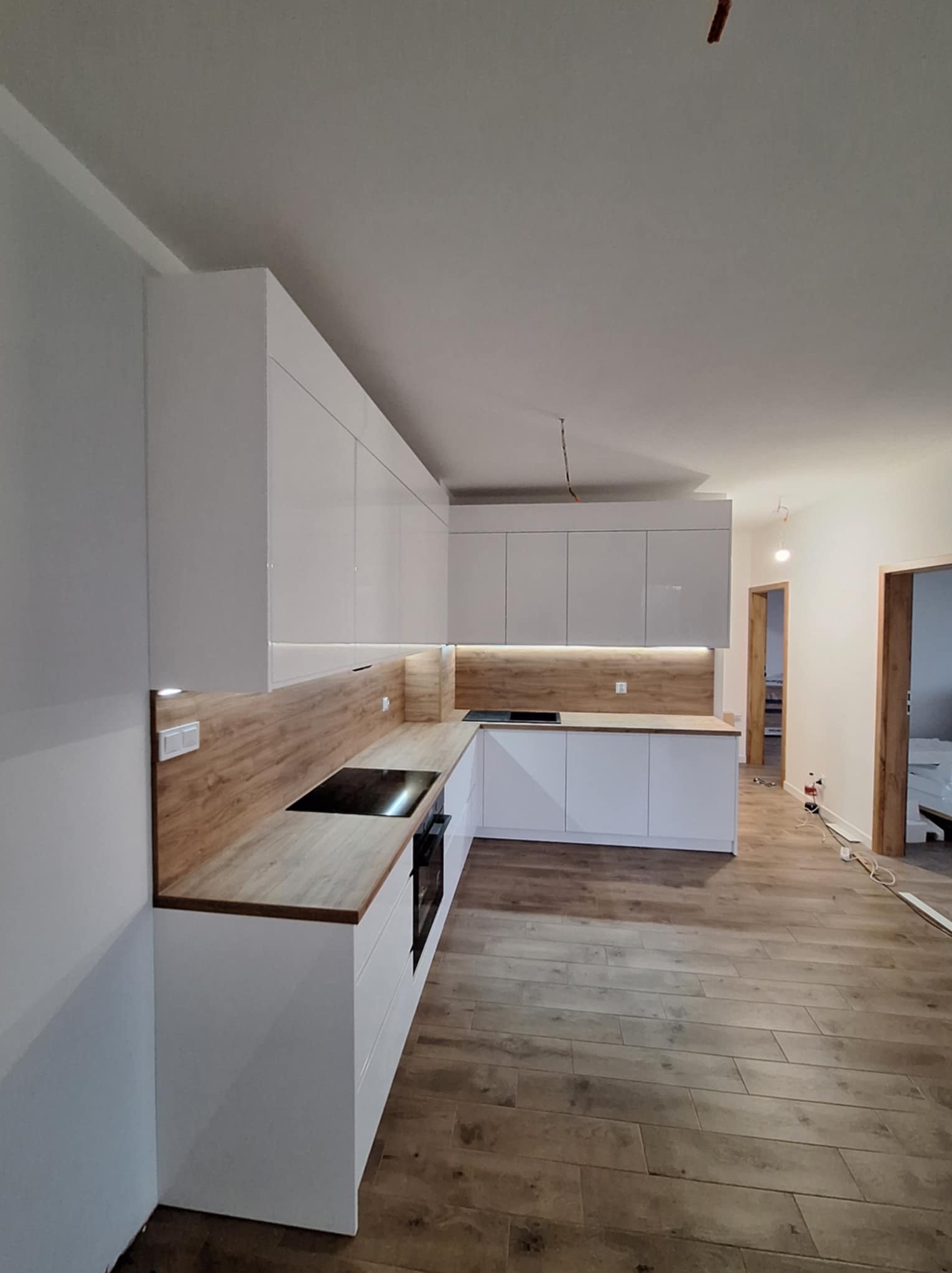 Nowoczesna kuchnia z białymi, lakierowanymi szafkami i drewnianym blatem, oświetlenie LED pod szafkami górnymi, widoczne niedokończone prace instalacyjne na suficie i podłodze.