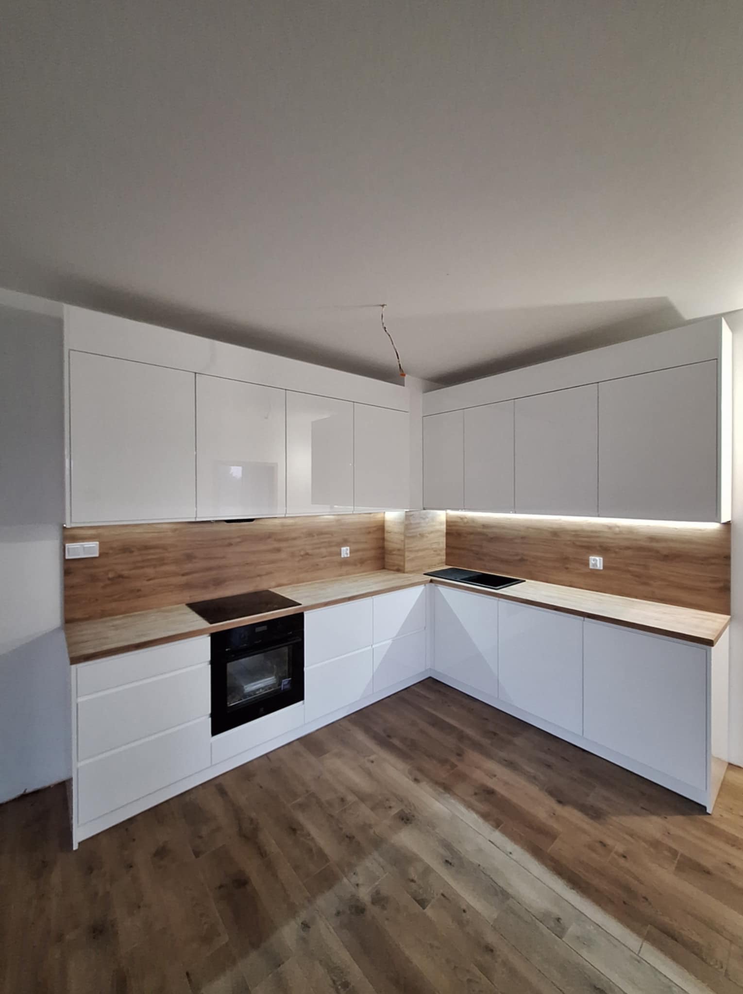 Nowoczesna, biała kuchnia z drewnianymi blatami i podświetleniem LED pod szafkami górnymi, widoczne niedokończone wykończenie ściany i wystające kable.