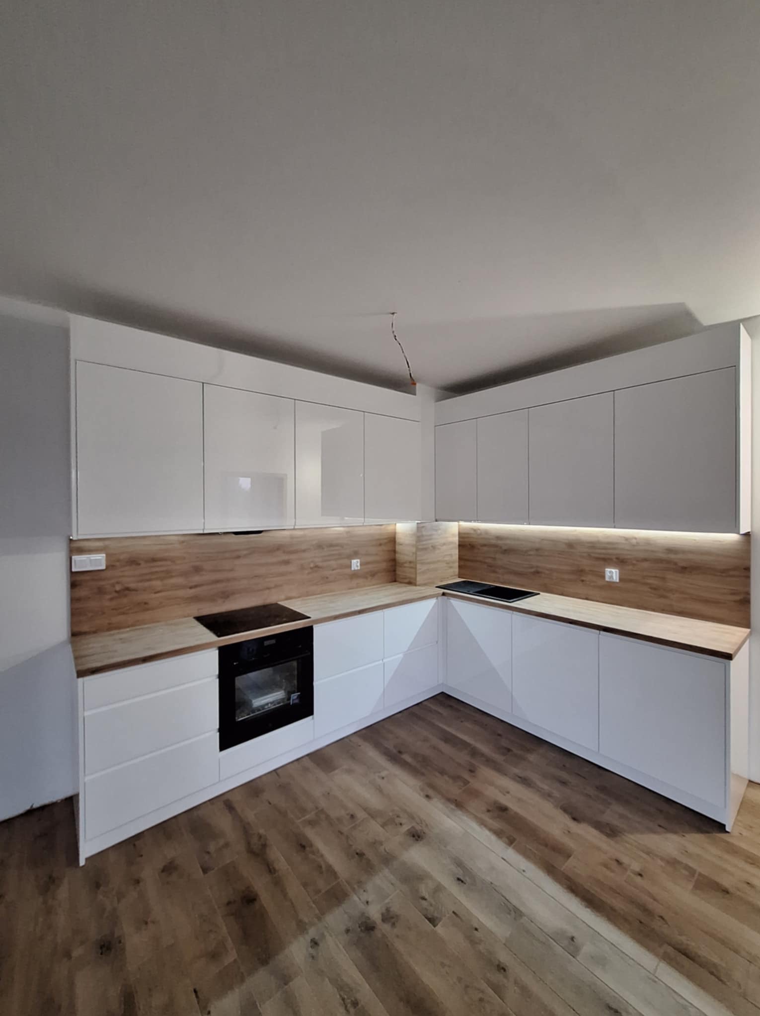Nowoczesna kuchnia z białymi, lakierowanymi frontami szafek i drewnianym blatem, oświetlona listwą LED pod szafkami wiszącymi, widok na narożnik i płytę grzewczą.