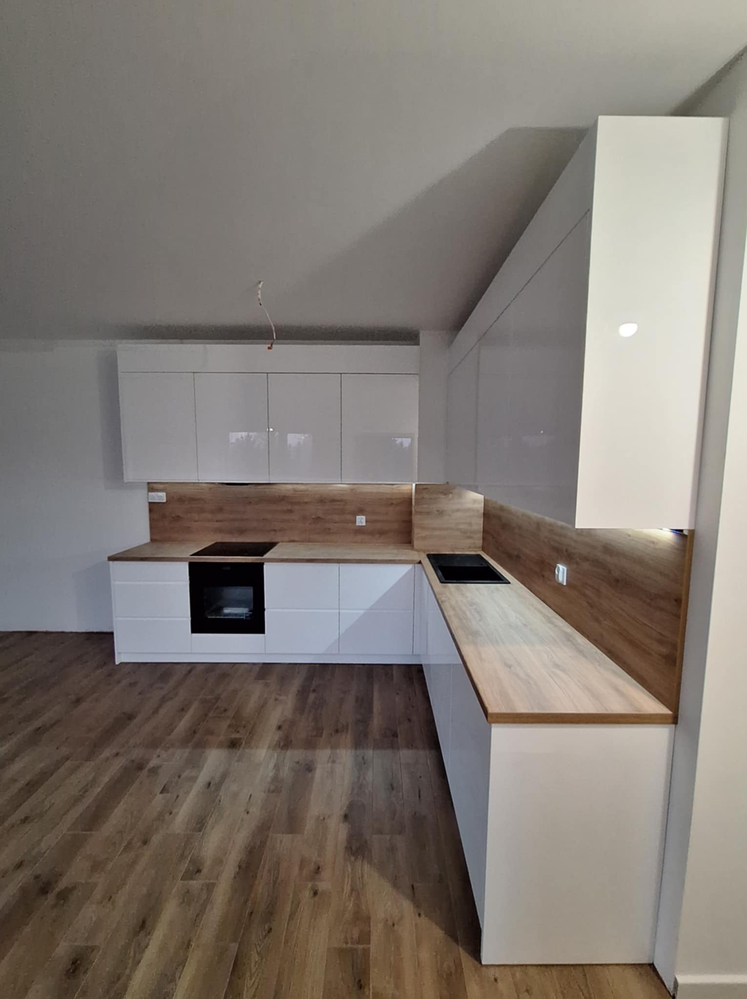 Nowoczesna, biała zabudowa kuchenna z drewnopodobnym blatem i panelem ściennym, widok na płytę indukcyjną i zlewozmywak, brązowe panele podłogowe.