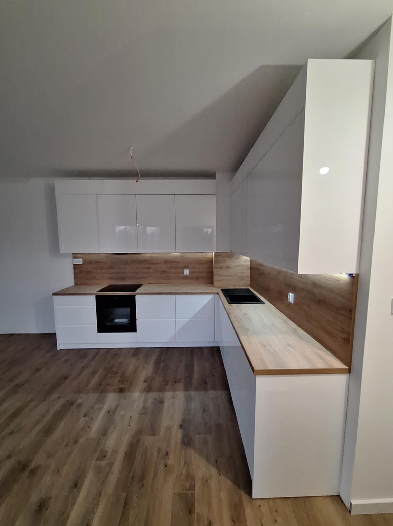 Nowoczesna, biała zabudowa kuchenna z drewnianym blatem i panelem ściennym w Kobiele Wielkie, z widoczną płytą indukcyjną i piekarnikiem. Na suficie widoczne niedokończone prace elektryczne.