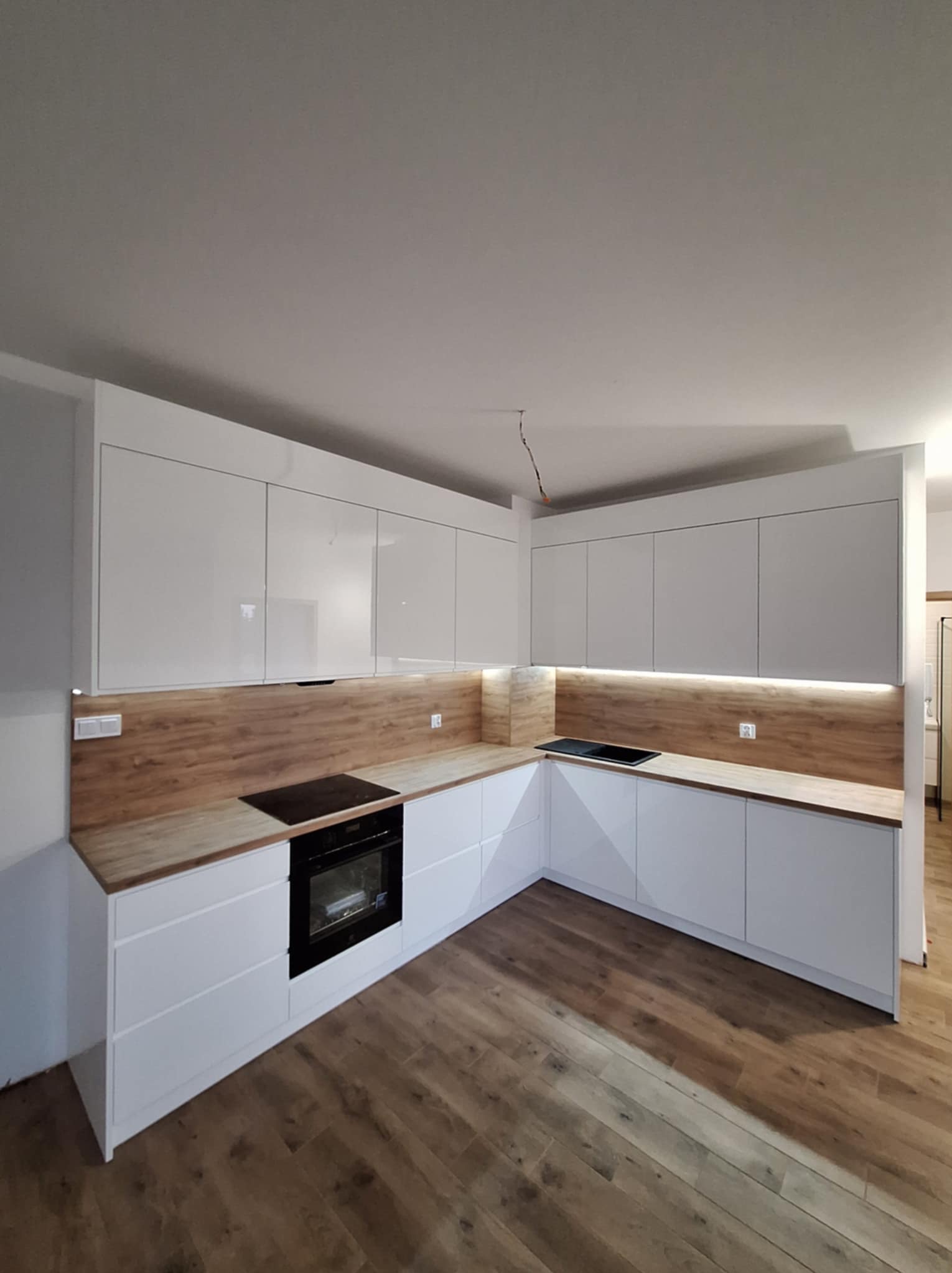 Nowoczesna kuchnia w kształcie litery L z białymi, błyszczącymi szafkami i drewnopodobnym blatem oraz oświetleniem LED pod szafkami wiszącymi, na podłodze panele imitujące drewno, widoczne...