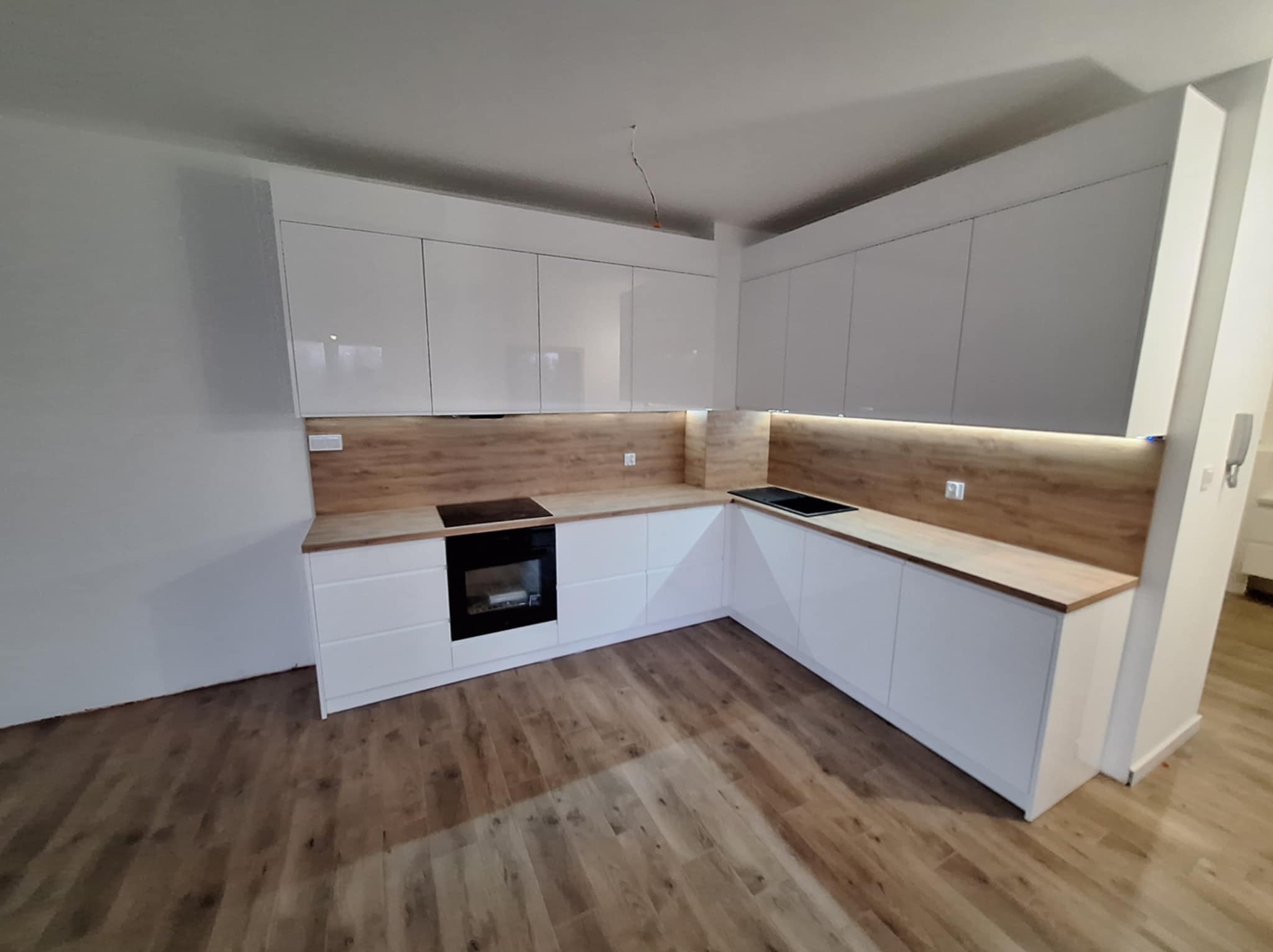 Nowoczesna kuchnia z białymi, lakierowanymi frontami szafek i drewnianym blatem, widoczna płyta indukcyjna i oświetlenie LED podszafkowe, surowy kabel zwisający z sufitu.