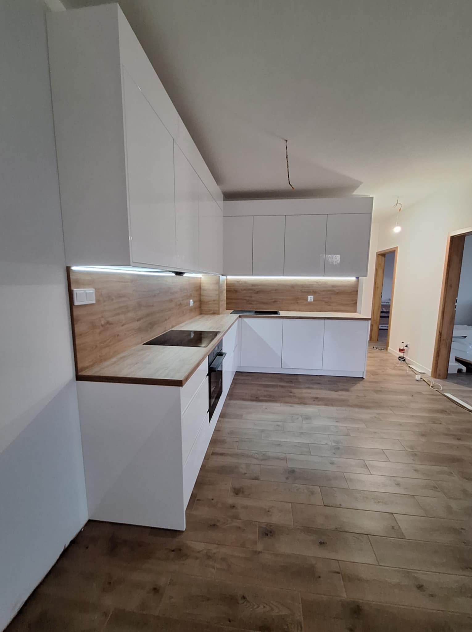 Nowoczesna kuchnia na wymiar z białymi, lakierowanymi frontami i drewnianym blatem, oświetlenie LED pod szafkami wiszącymi, widok na drewnopodobną podłogę i otwarte drzwi do innych pomieszczeń.