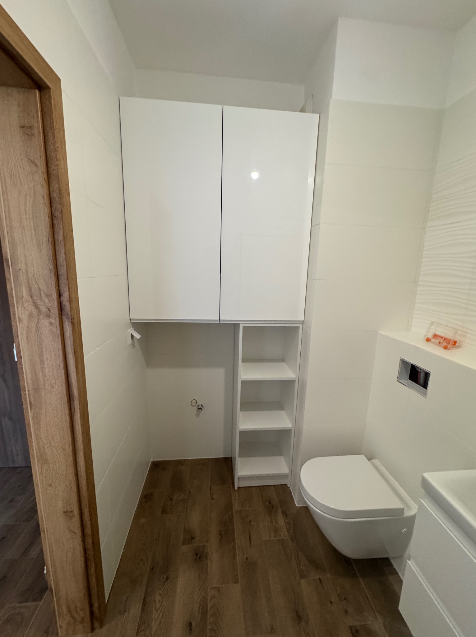 Nowoczesna łazienka z białymi, wiszącymi szafkami, otwartymi półkami i podwieszaną toaletą na tle płytek imitujących drewno.