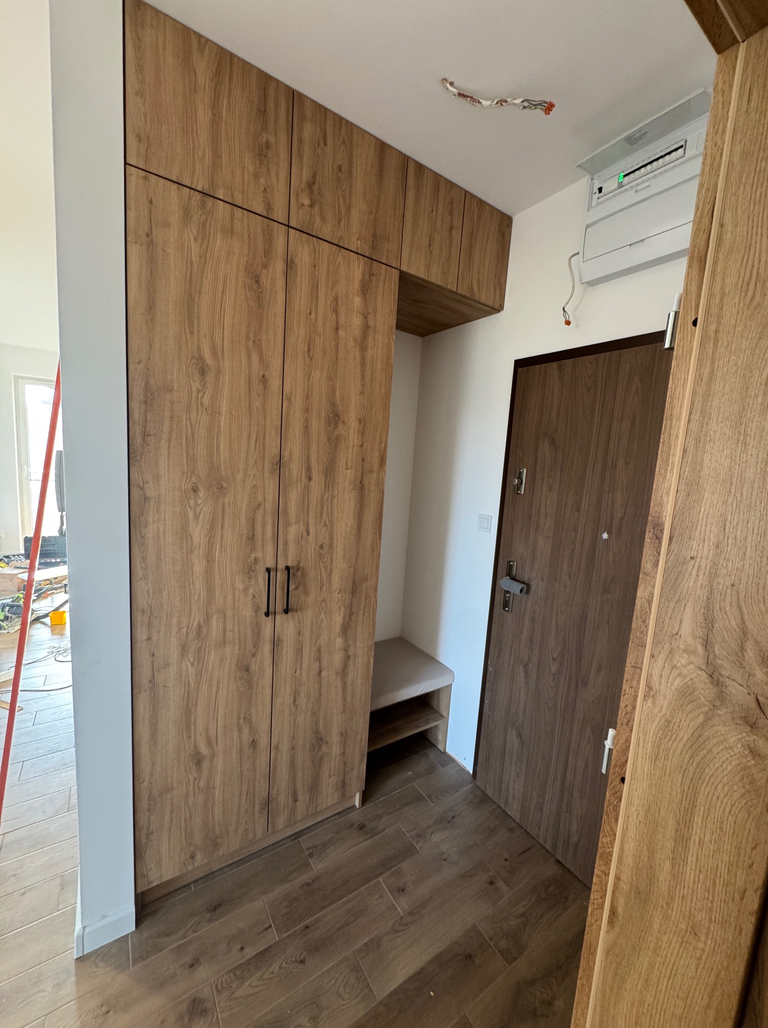 Przedpokój z zabudową: szafa z drewnopodobnymi frontami i siedziskiem, dopasowana do drzwi wejściowych, widoczne niedokończone prace instalacyjne na suficie.