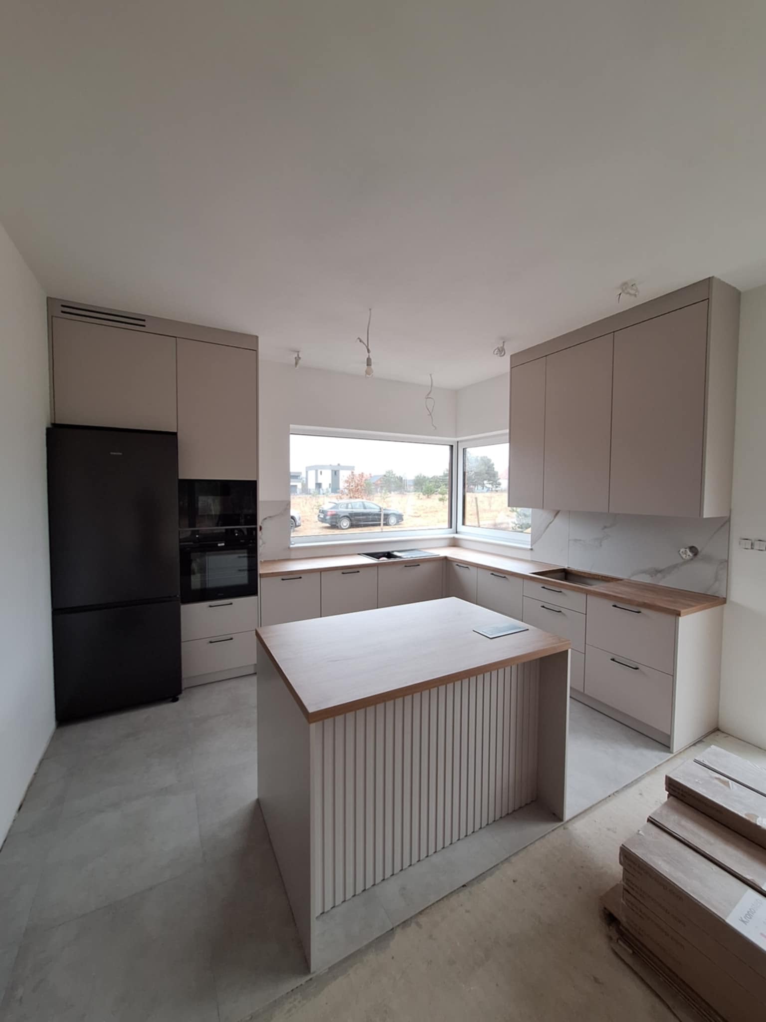 Jasna kuchnia z wyspą, fronty szafek w kolorze beżowym, czarny sprzęt AGD, częściowo wykończona podłoga, widoczne niedociągnięcia wykończeniowe.