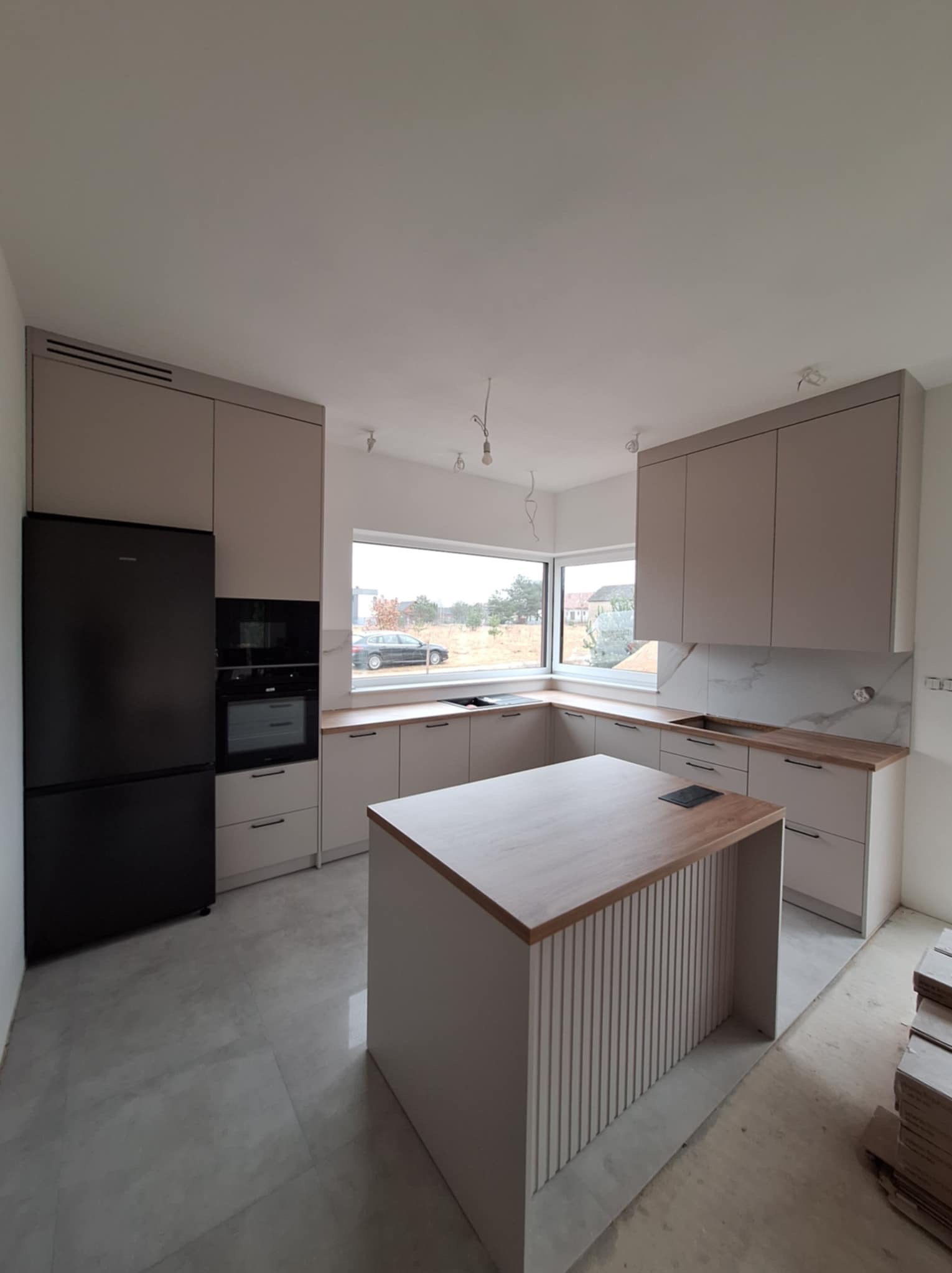 Beżowa zabudowa kuchenna z czarną lodówką i wyspą z ryflowanym frontem, widok na okno z szarą zabudową na zewnątrz, na podłodze szare płytki, widoczne niedokończone prace wykończeniowe.