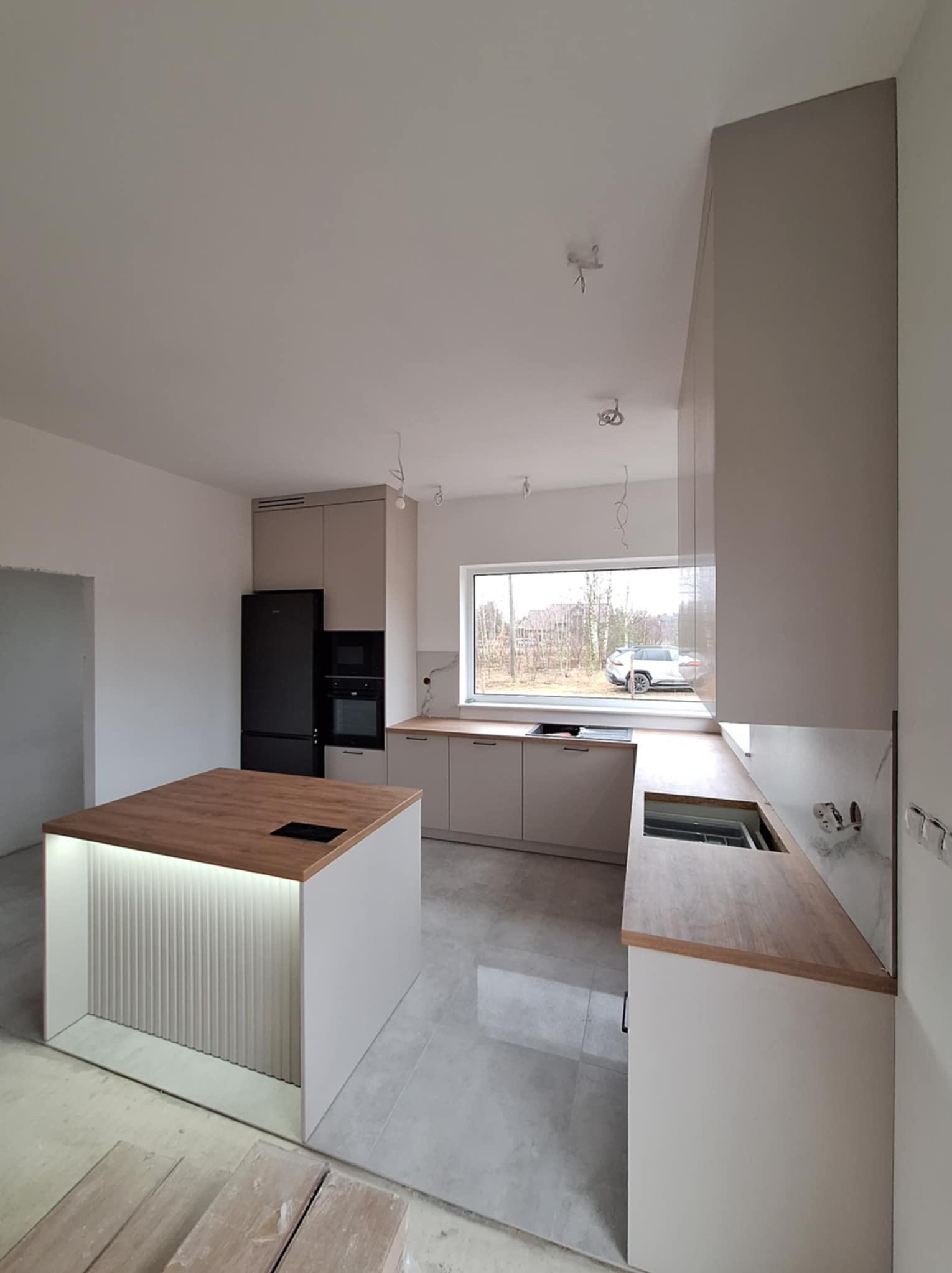 Nowoczesna kuchnia w trakcie montażu: wyspa z podświetleniem LED, drewniany blat, szare szafki i czarna lodówka w surowym wnętrzu.