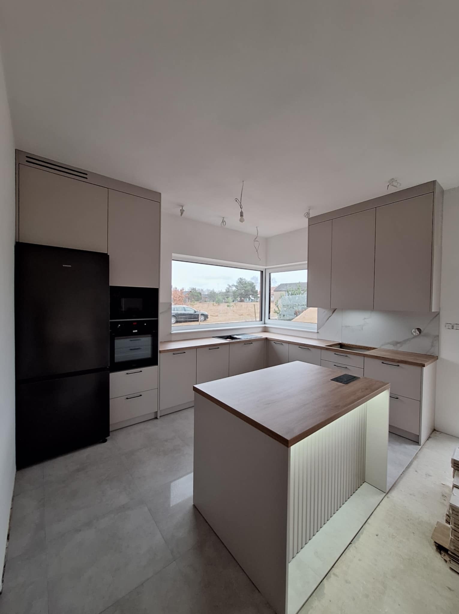 Jasna kuchnia z wyspą i czarną lodówką, widok na okno z zabudowy kuchennej, podświetlana wyspa kuchenna z dekoracyjnymi panelami pionowymi, szare płytki podłogowe.