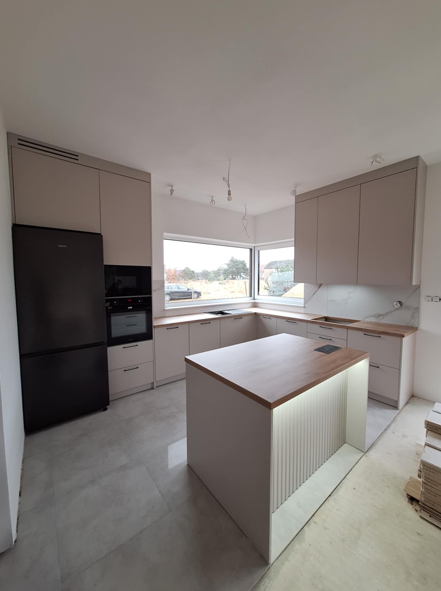 Jasna kuchnia z wyspą, czarnym sprzętem AGD i marmurowym backsplashiem, widok na zewnątrz przez okno, surowy stan wykończenia podłogi.