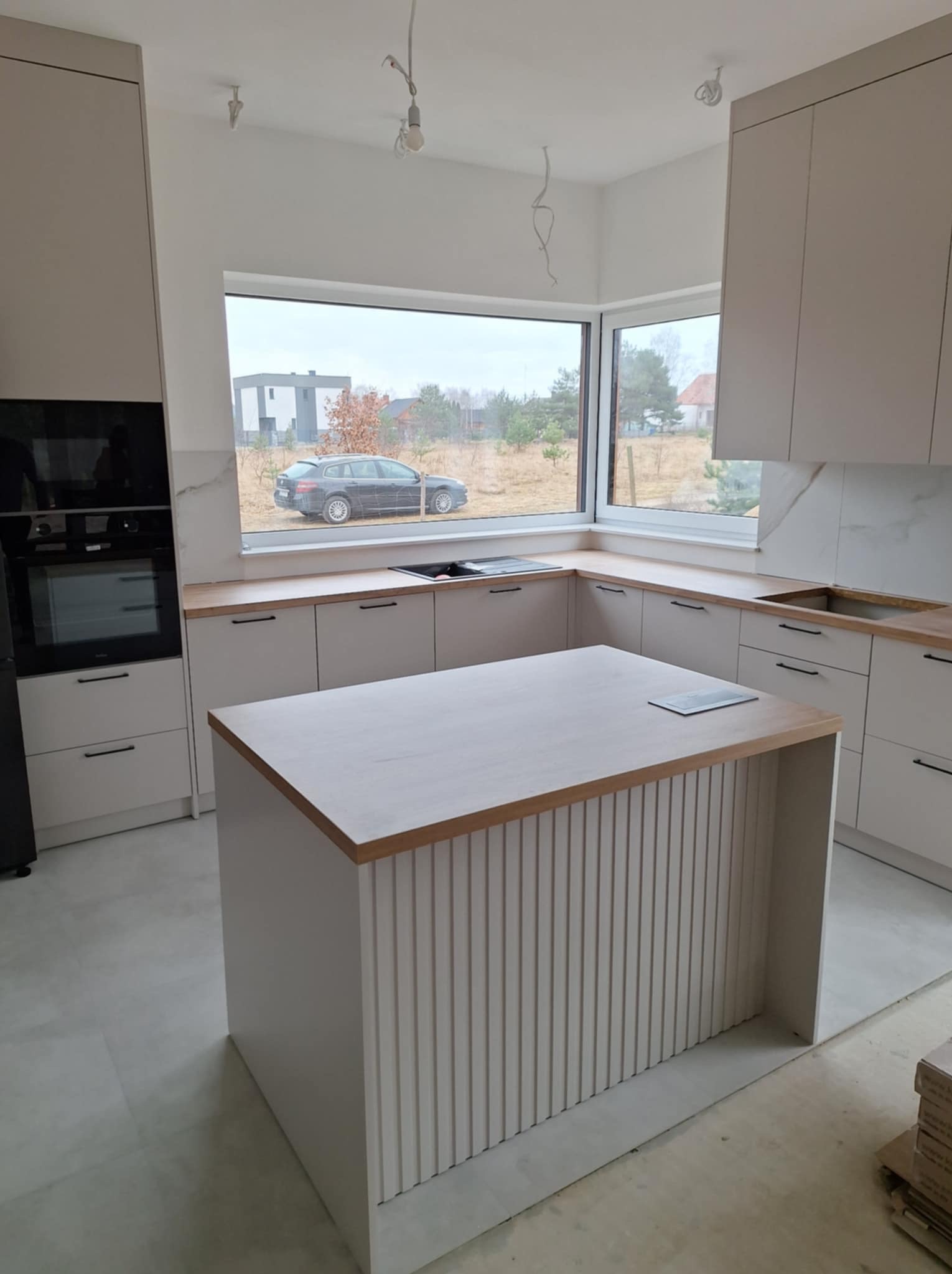 Jasna kuchnia z wyspą i drewnianymi blatami, widok na szare niebo i samochód za oknem, niedokończone wykończenie podłogi.