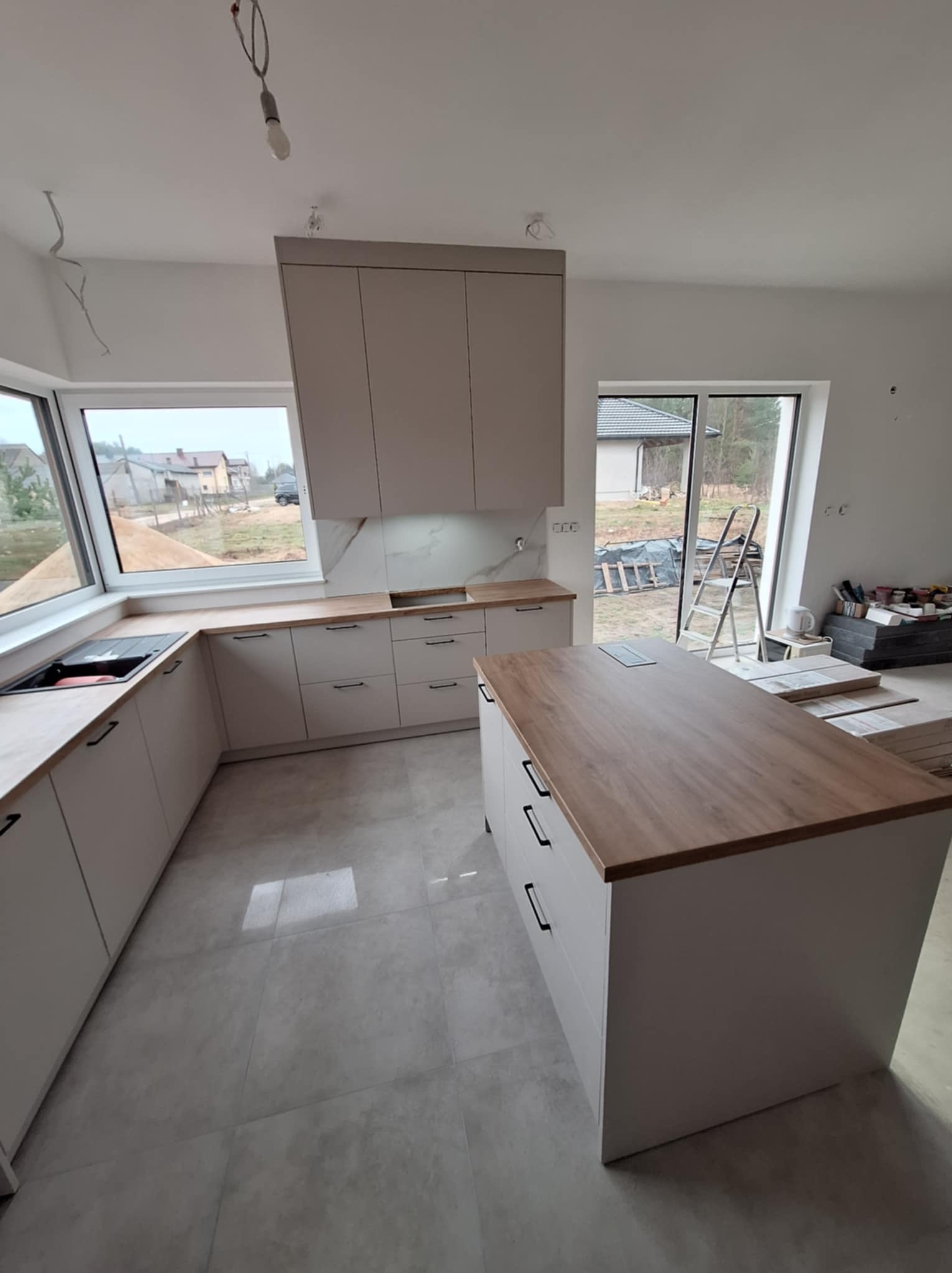 Jasna, nowoczesna kuchnia z wyspą, widoczna w trakcie montażu; surowe wykończenie wnętrza, brak lampy sufitowej, okno narożne z widokiem na zabudowę jednorodzinną.