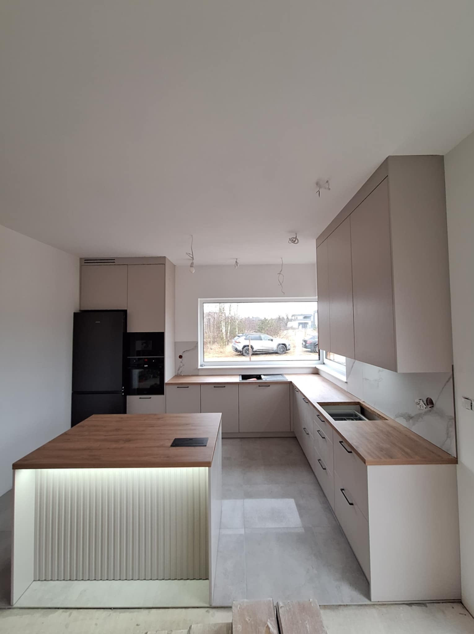 Nowoczesna kuchnia w trakcie montażu: beżowe szafki z drewnianymi blatami, wyspa z oświetleniem LED, czarna lodówka i piekarnik, widoczne niedokończone wykończenia ścian i sufitu.