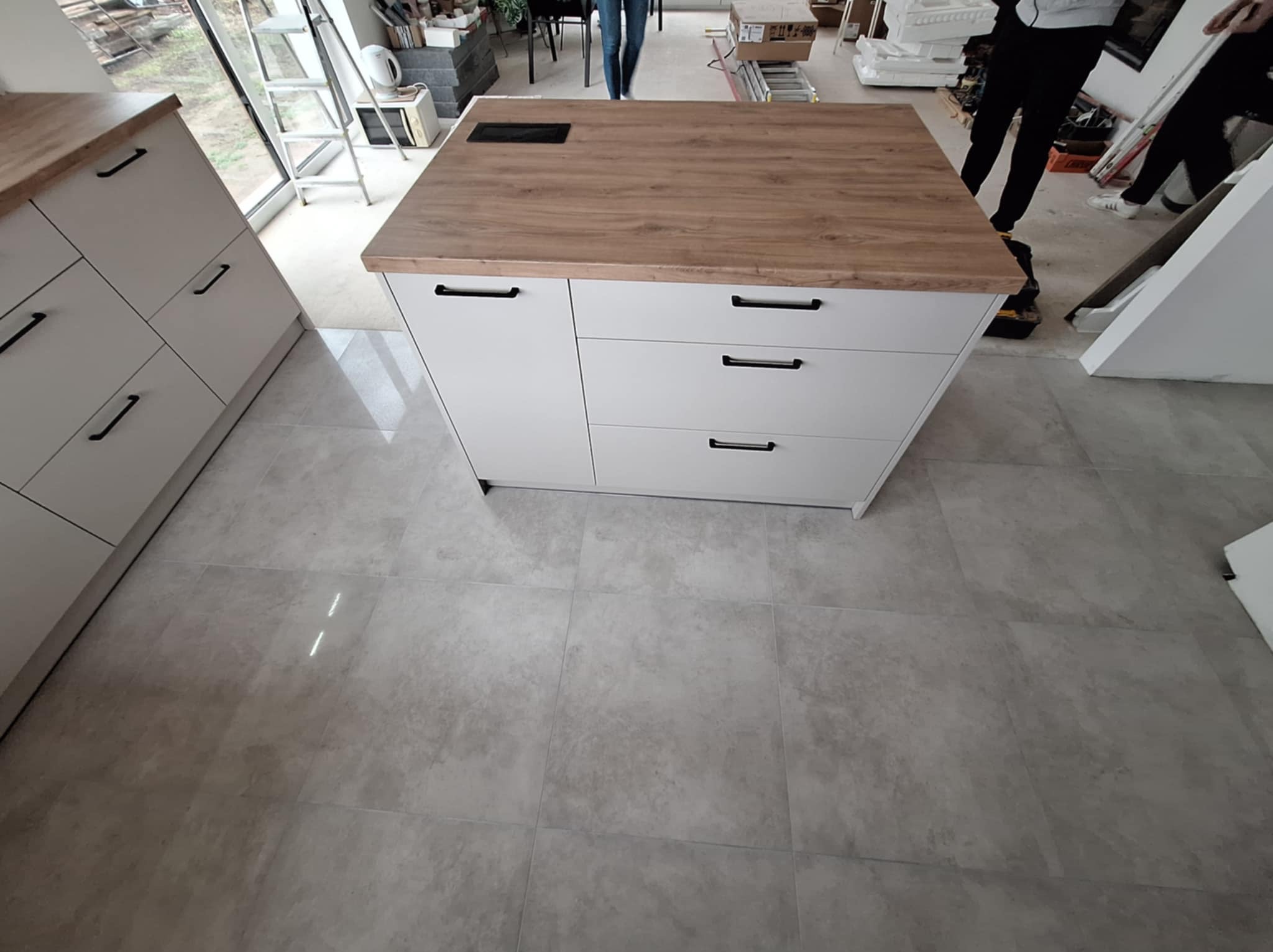 Wyspa kuchenna z białymi frontami i blatem imitującym drewno, wyposażona w czarne, poziome uchwyty, widoczna w trakcie montażu w pomieszczeniu z szarą podłogą z płytek.