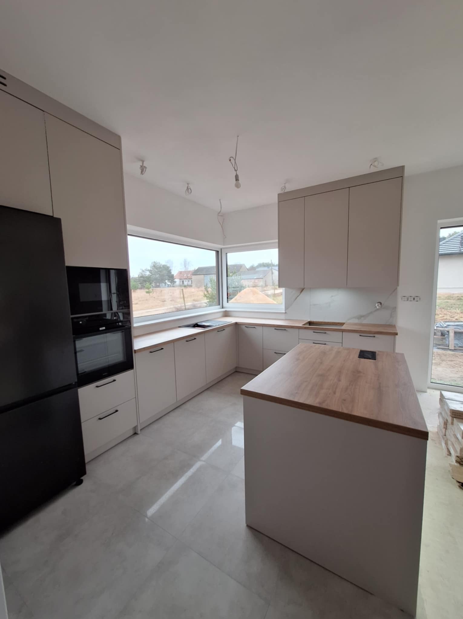 Nowoczesna kuchnia w jasnych odcieniach z wyspą i czarną lodówką w zabudowie, widok na okno z widokiem na budowę.