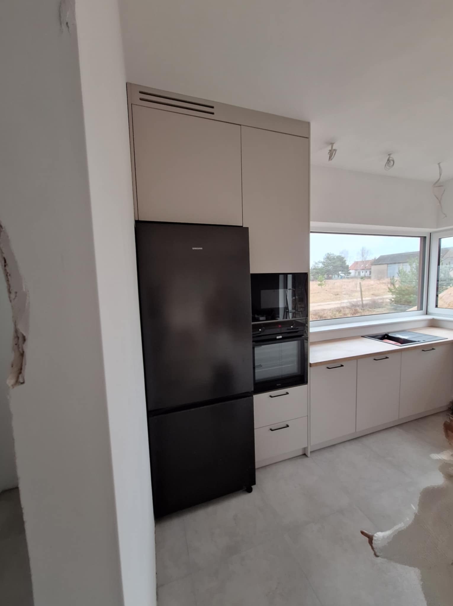 Jasna kuchnia z czarną lodówką i piekarnikiem w zabudowie, widok na okno z drewnianym blatem, surowy stan wykończenia ściany po lewej stronie.