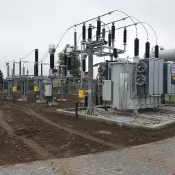 Zewnętrzna stacja transformatorowa z licznymi izolatorami i wentylatorami chłodzącymi, otoczona ziemią i kostką brukową.