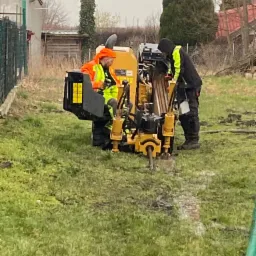 Dwóch pracowników w odblaskowych ubraniach obsługuje żółtą wiertnicę na trawniku, widoczne ślady wiercenia w ziemi, w tle ogrodzenie i budynki.