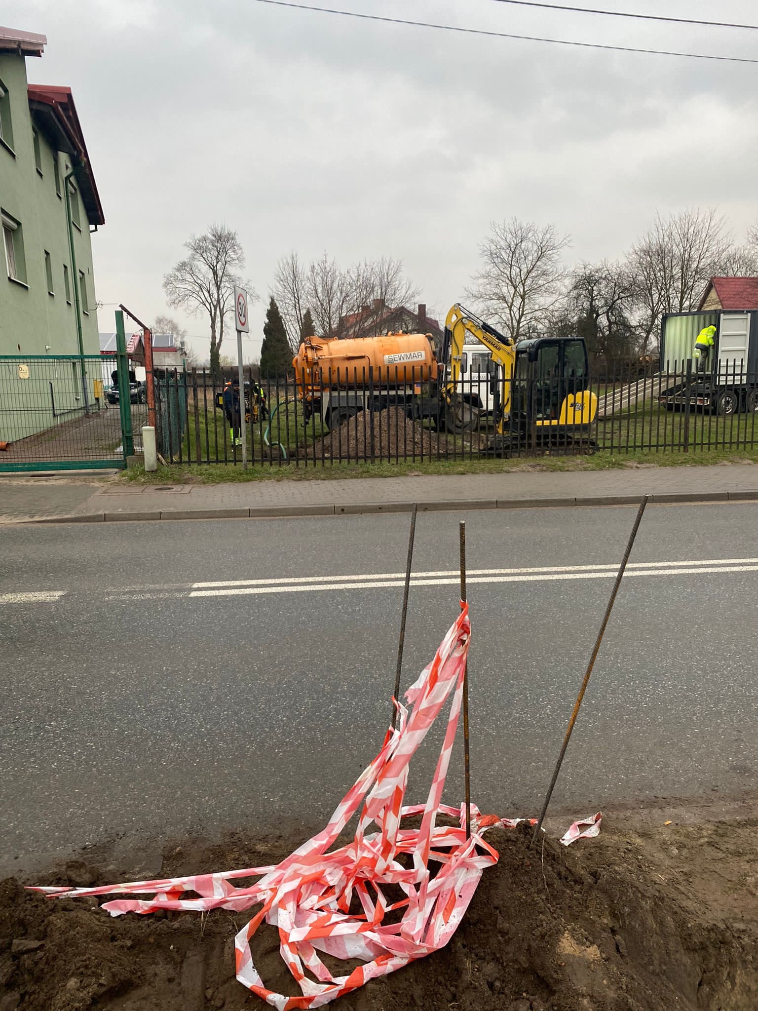 Prace ziemne przy drodze: widoczny wykop zabezpieczony taśmą ostrzegawczą, w tle żółta cysterna asenizacyjna i mała koparka Yanmar, sugerujące prace hydrauliczne lub związane z wierceniem studni.