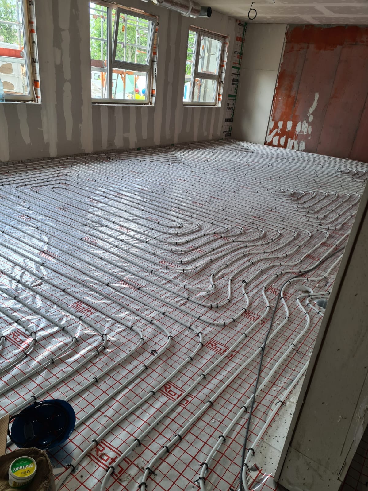 Białe rury ogrzewania podłogowego ułożone spiralnie na folii z czerwoną kratką w surowym pomieszczeniu z oknami i częściowo otynkowanymi ścianami.