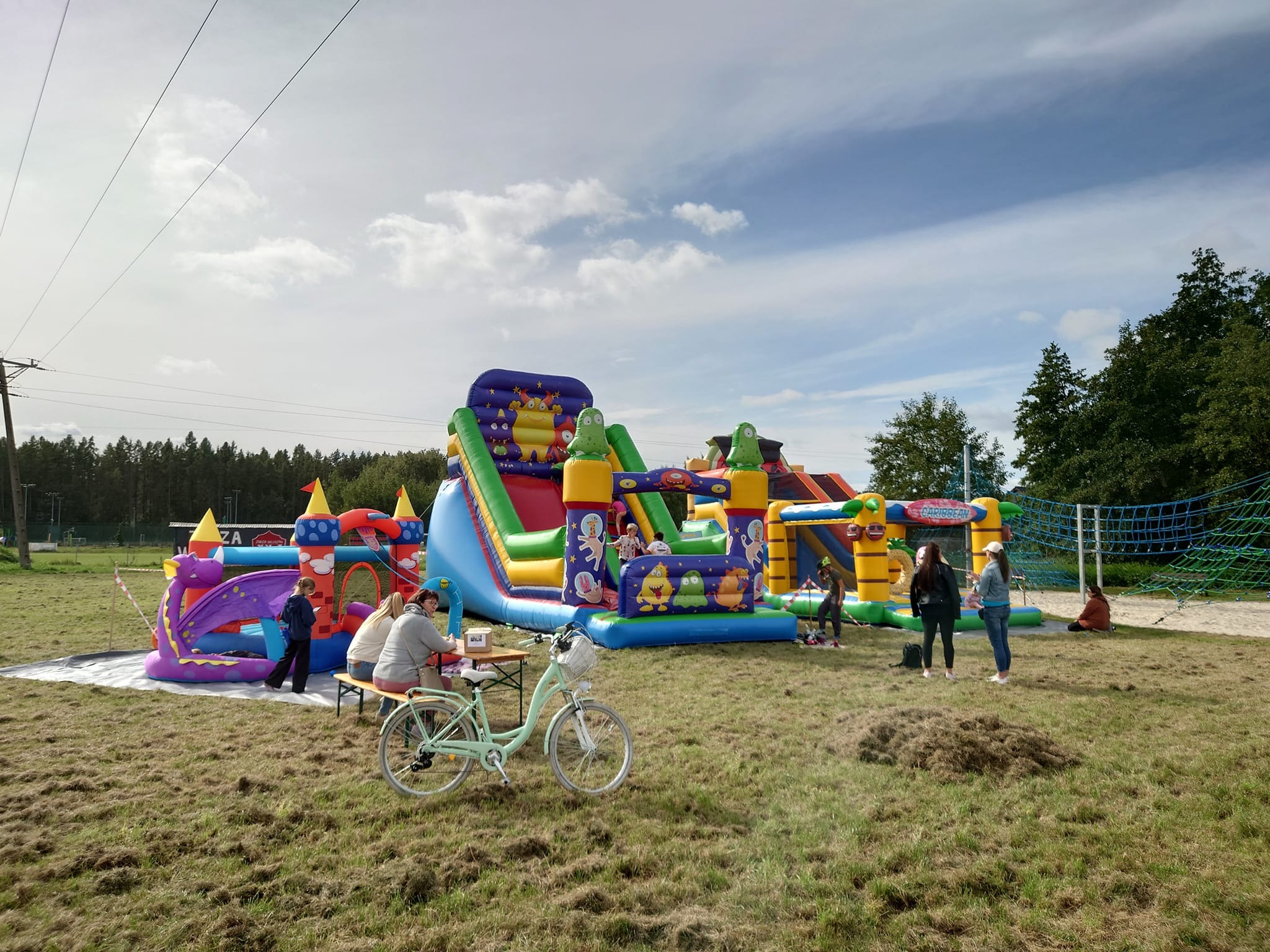 Rozległy plac zabaw na świeżym powietrzu z dmuchanymi zamkami i zjeżdżalniami, na pierwszym planie jasnoturkusowy rower oparty o stolik z siedzącymi osobami, w tle bawiące się dzieci.