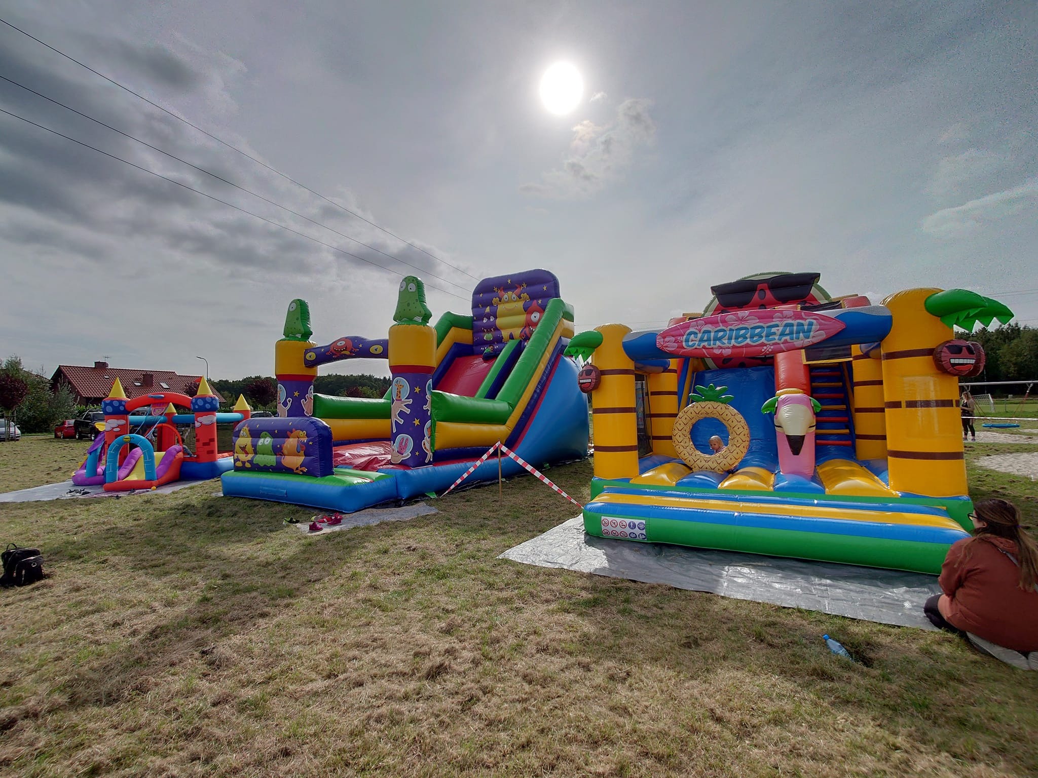 Kilka kolorowych, dmuchanych zamków na trawiastym terenie, w tle budynek z czerwoną dachówką i pochmurne niebo z widocznym słońcem.