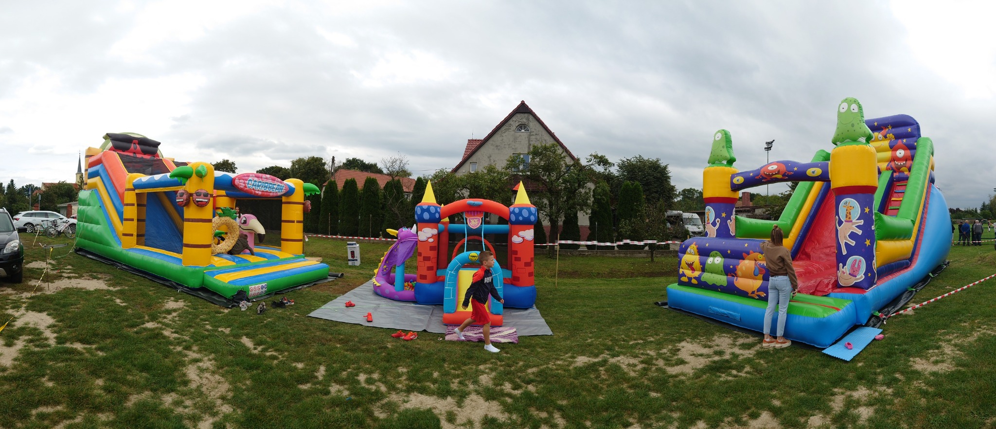 Panorama trzech kolorowych, dmuchanych zamków na trawiastym terenie z dziećmi, w tle dom i pochmurne niebo.