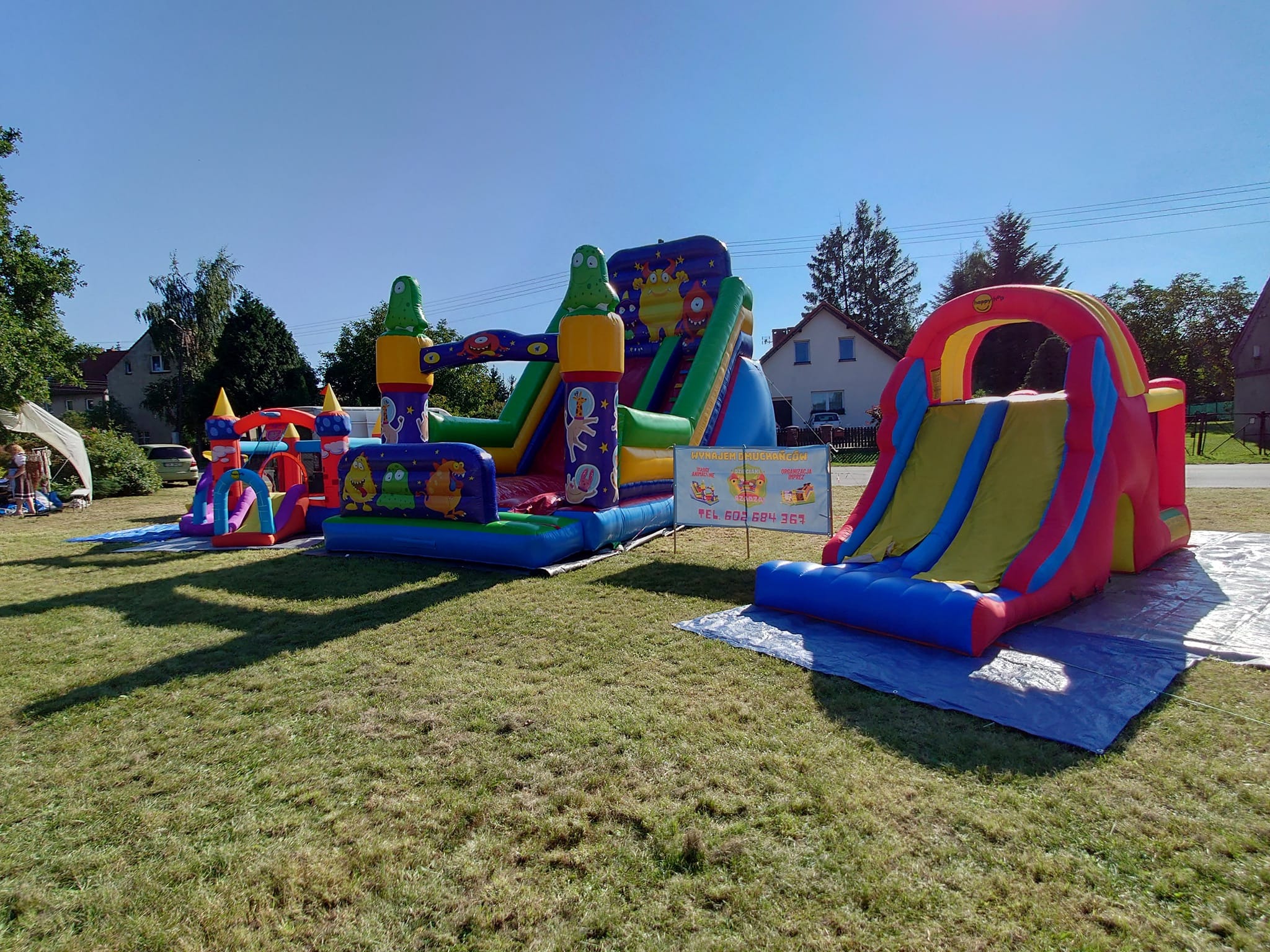 Kompleks dmuchańców na trawie: zjeżdżalnie, zamki, kolorowe postacie, widoczny baner reklamowy z numerem telefonu.