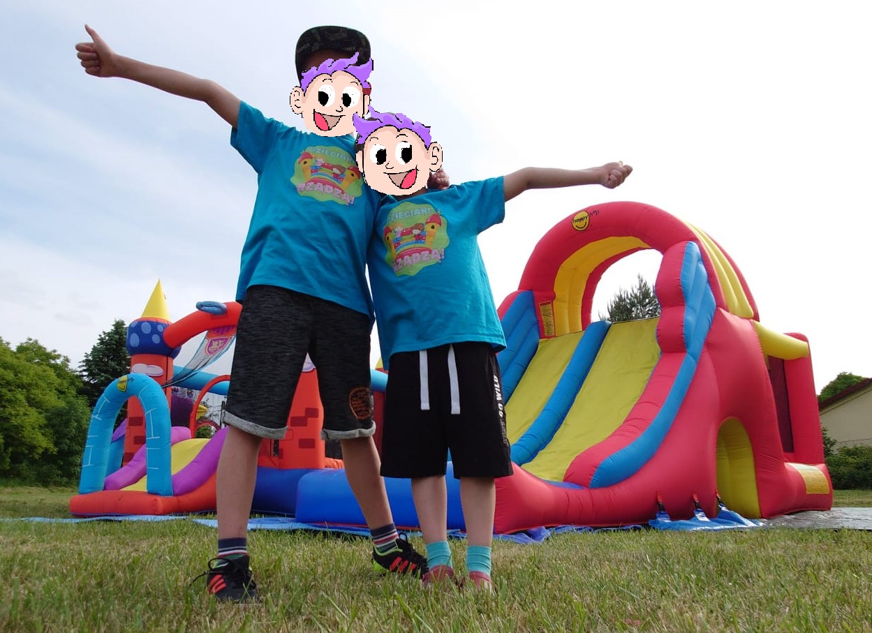 Dwaj chłopcy w niebieskich koszulkach z grafiką stoją przed kolorowym dmuchanym zamkiem ze zjeżdżalnią na trawniku.