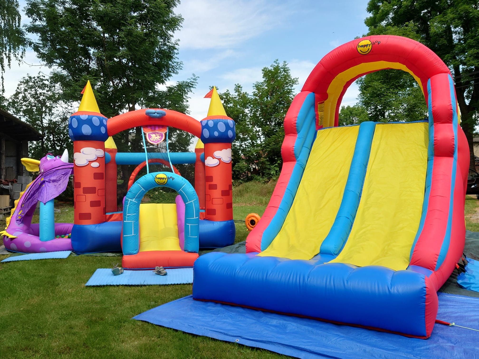 Rozłożone na trawie, kolorowe dmuchańce z motywem zamku i zjeżdżalni, marki Happy Hop, na tle drzew i częściowo widocznego budynku.