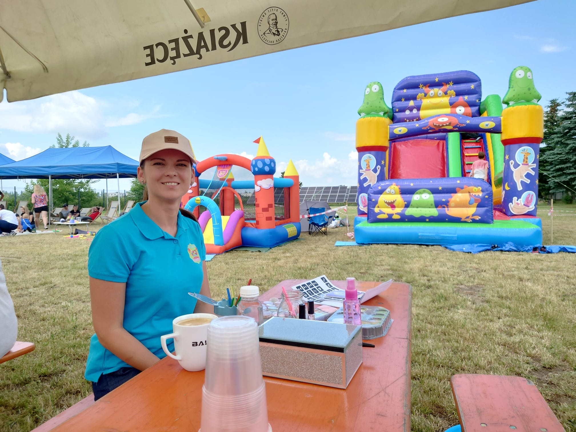 Uśmiechnięta kobieta w turkusowej koszulce siedzi przy stole z kawą i akcesoriami do malowania twarzy, w tle dmuchane zamki i bawiące się dzieci.