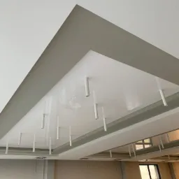 Biały, podwieszany sufit z oświetleniem punktowym w kształcie cylindrów, widok z dołu, szara obudowa sufitu i kratki wentylacyjne.