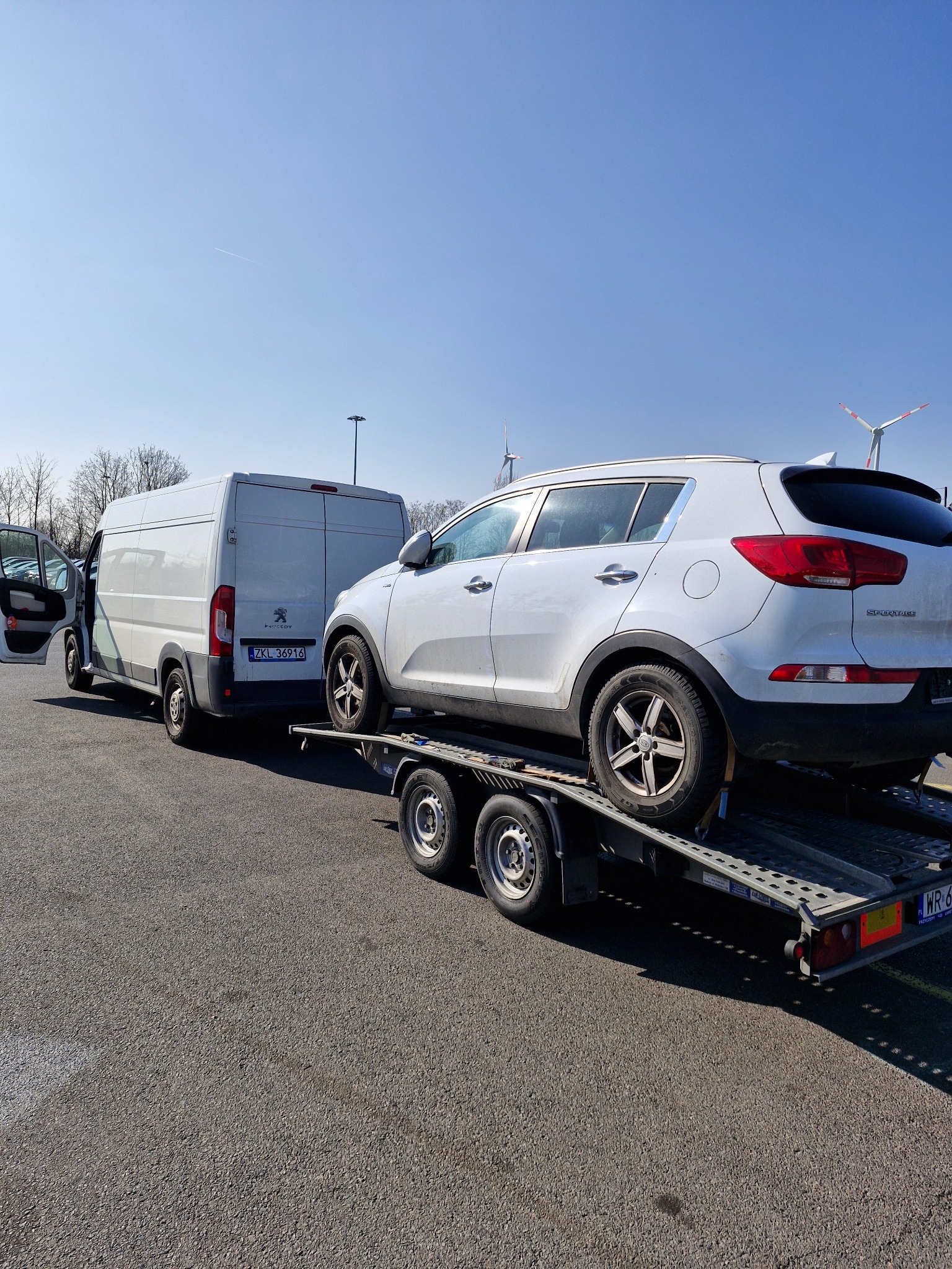 Biały samochód Kia Sportage zabezpieczony pasami na lawecie, ciągniętej przez białego dostawczego Peugeota, na tle wiatraków i błękitnego nieba.