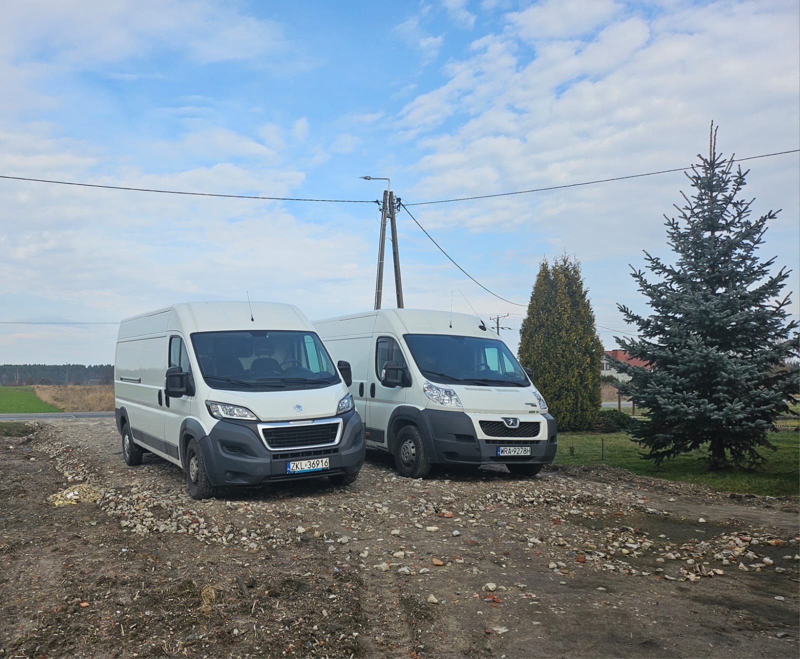 Dwa białe busy Peugeot zaparkowane na żwirowym podłożu w wiejskim otoczeniu, z widocznymi numerami rejestracyjnymi ZKL 36916 i WRA 9278H, na tle błękitnego nieba z chmurami i drzew iglastych.