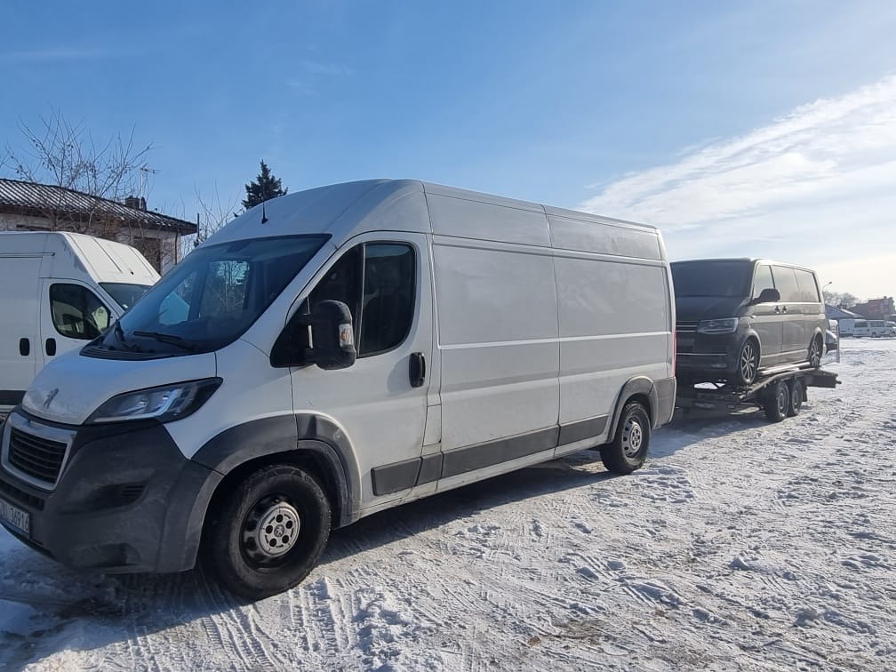 Biały bus z przyczepą, na której przewożony jest ciemny samochód osobowy, zimowy krajobraz.