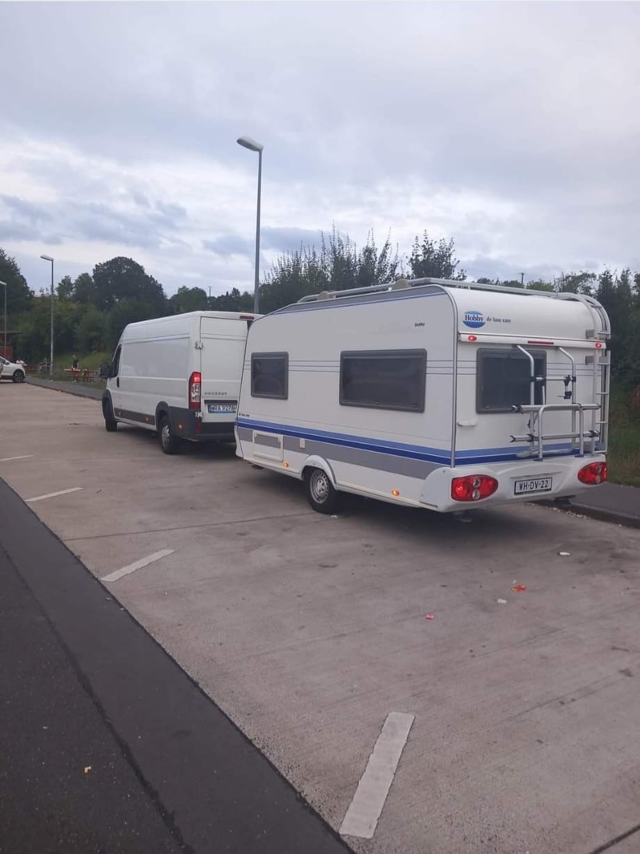 Biały bus ciągnie przyczepę kempingową marki Hobby na parkingu, widoczne tablice rejestracyjne pojazdów.