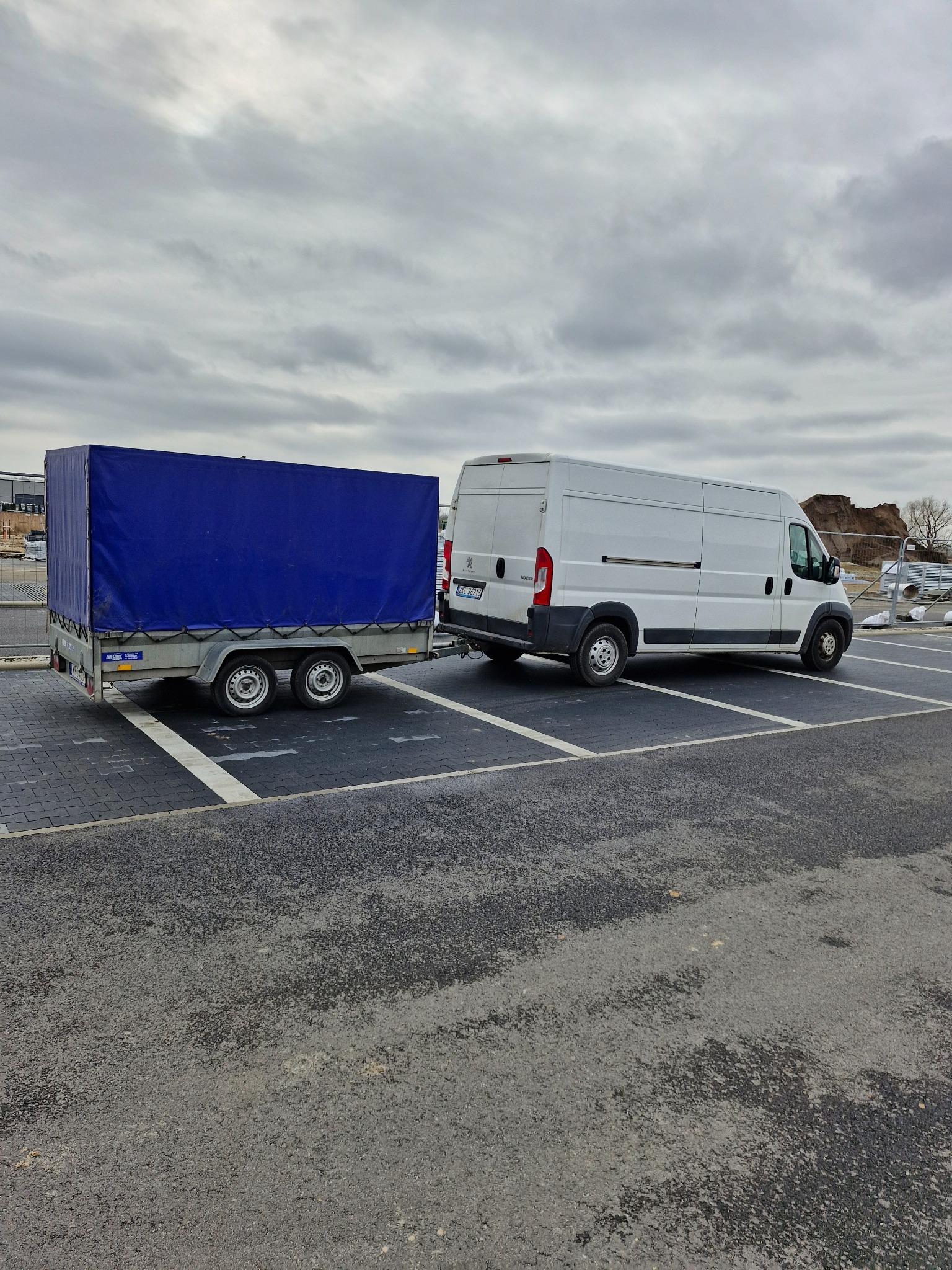 Biały van z niebieską przyczepą na parkingu, pochmurne niebo w tle.