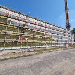 Adbud usługi ogólnobudowlane - Prace przy ocieplaniu ściany budynku przemysłowego z wysokim kominem w tle, widoczne rusztowania i pracownicy montujący izolację termiczną.
