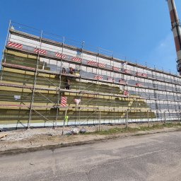 Adbud usługi ogólnobudowlane - Ocieplanie elewacji budynku przemysłowego z widocznymi rusztowaniami, izolacją z wełny mineralnej i pracownikami w trakcie montażu, w tle komin przemysłowy.