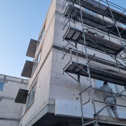 Adbud usługi ogólnobudowlane - Budynek wielorodzinny w trakcie prac wykończeniowych elewacji, widoczne rusztowania i pracownik w szarym kombinezonie montujący izolację termiczną na ścianie z betonu komórkowego.