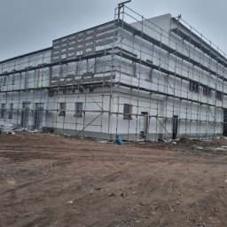 Adbud usługi ogólnobudowlane - Budynek w trakcie prac elewacyjnych, otoczony rusztowaniami, z widocznymi workami materiałów budowlanych i błotnistym terenem wokół.