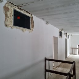 Adbud usługi ogólnobudowlane - Wnętrze remontowanego poddasza z oknami w trakcie montażu, uszczelnionymi pianką montażową, widoczne drewniane belki stropowe i fragment balustrady.