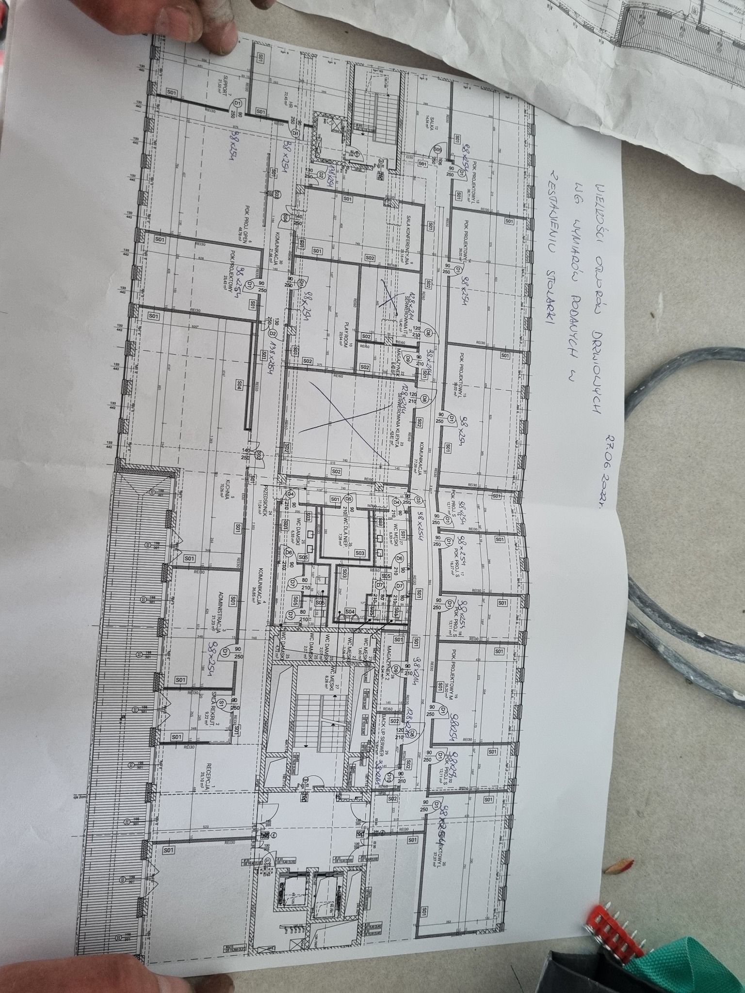 Rzut architektoniczny budynku wielorodzinnego z naniesionymi wymiarami i opisem pomieszczeń, częściowo zasłonięty dłońmi.