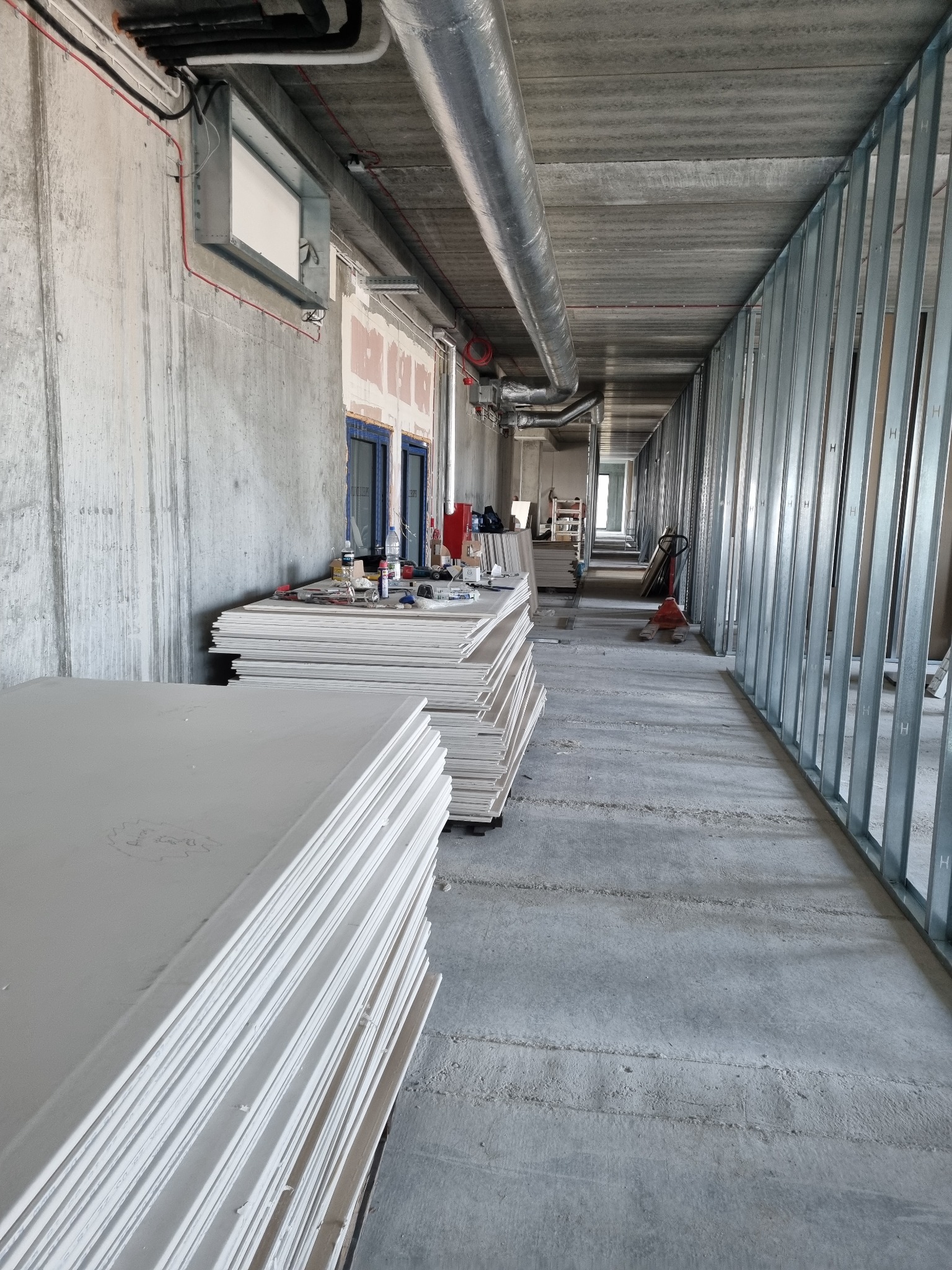 Wnętrze budynku w trakcie prac wykończeniowych; widoczne płyty gipsowo-kartonowe, metalowe profile konstrukcyjne ścian działowych, instalacje pod sufitem, narzędzia pracy.