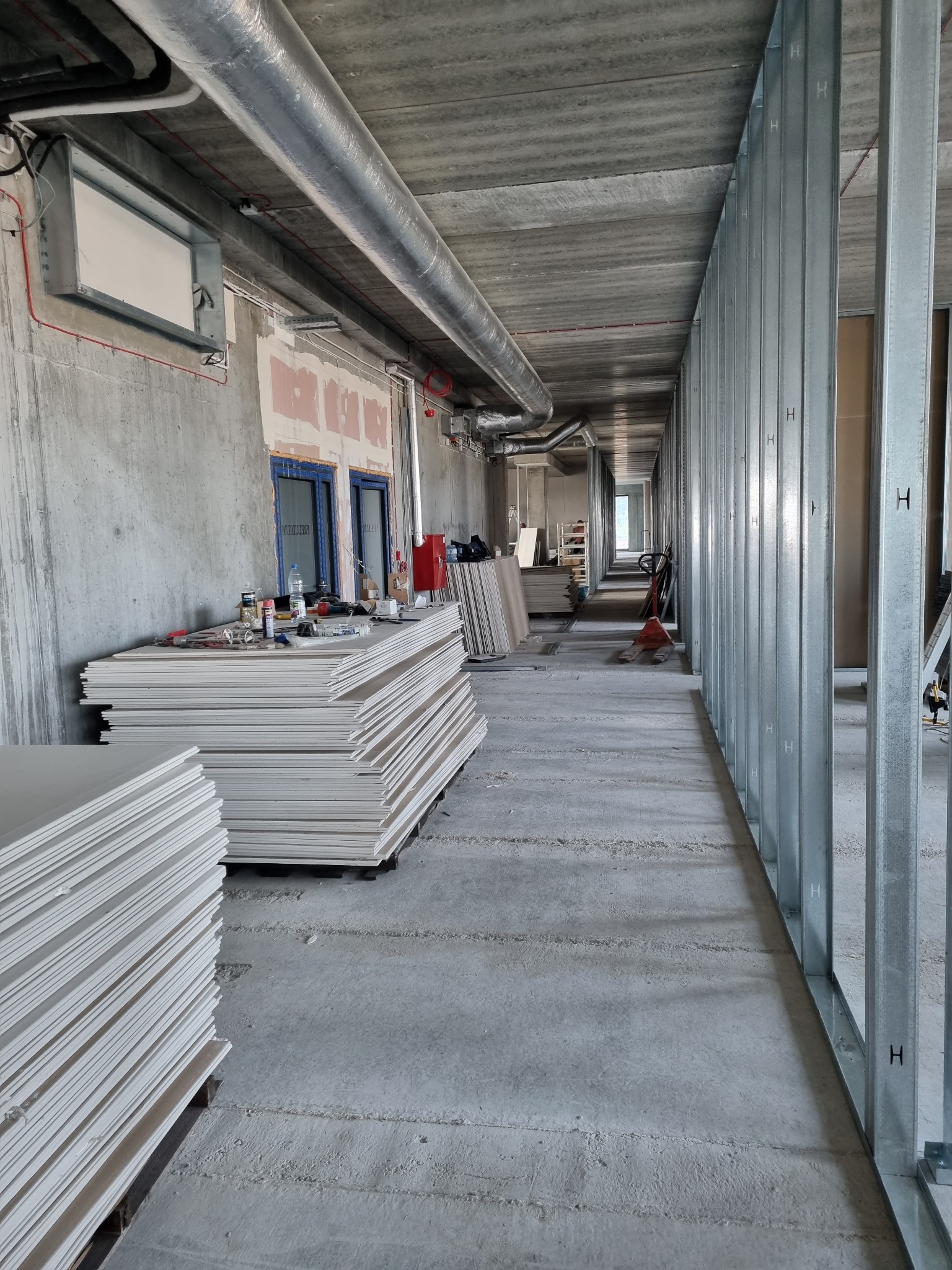 Wnętrze budynku w trakcie prac wykończeniowych: stosy płyt gipsowo-kartonowych, metalowe profile ścian działowych, instalacje pod sufitem, surowy beton.