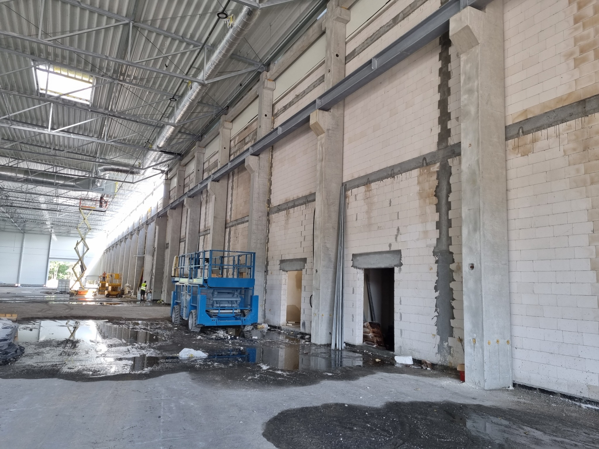 Wnętrze hali w trakcie budowy z widoczną ścianą z białych bloczków, stalowymi belkami pod stropem, niebieskim podnośnikiem nożycowym i kałużami na podłodze.